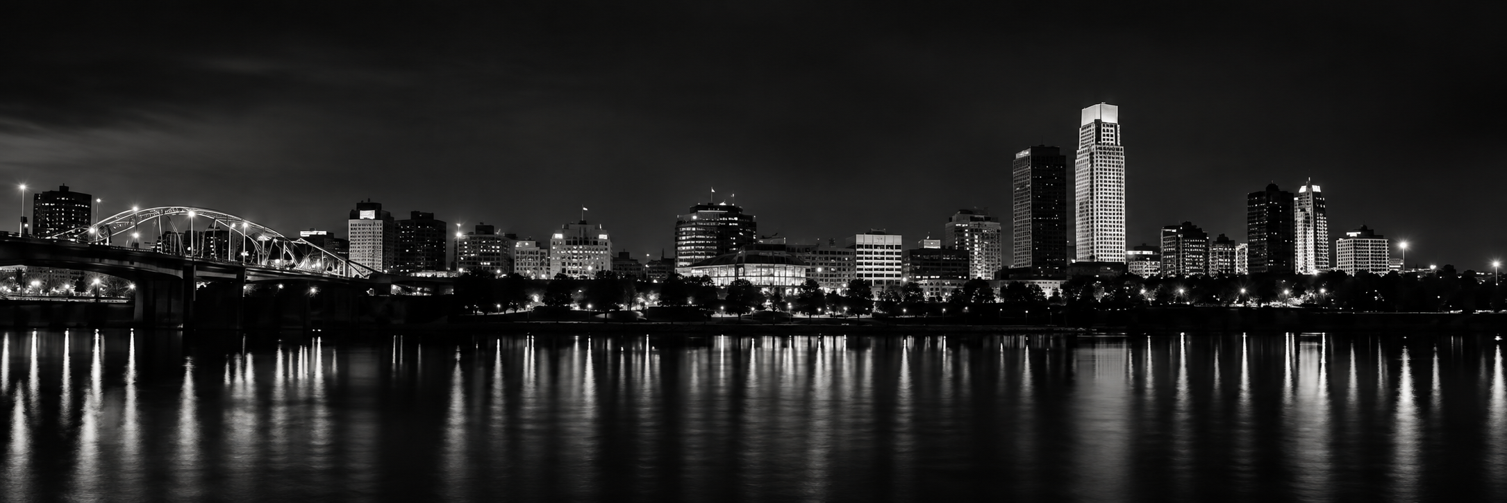 Nighttime city skyline with illuminated buildings and reflections on the water in the foreground.