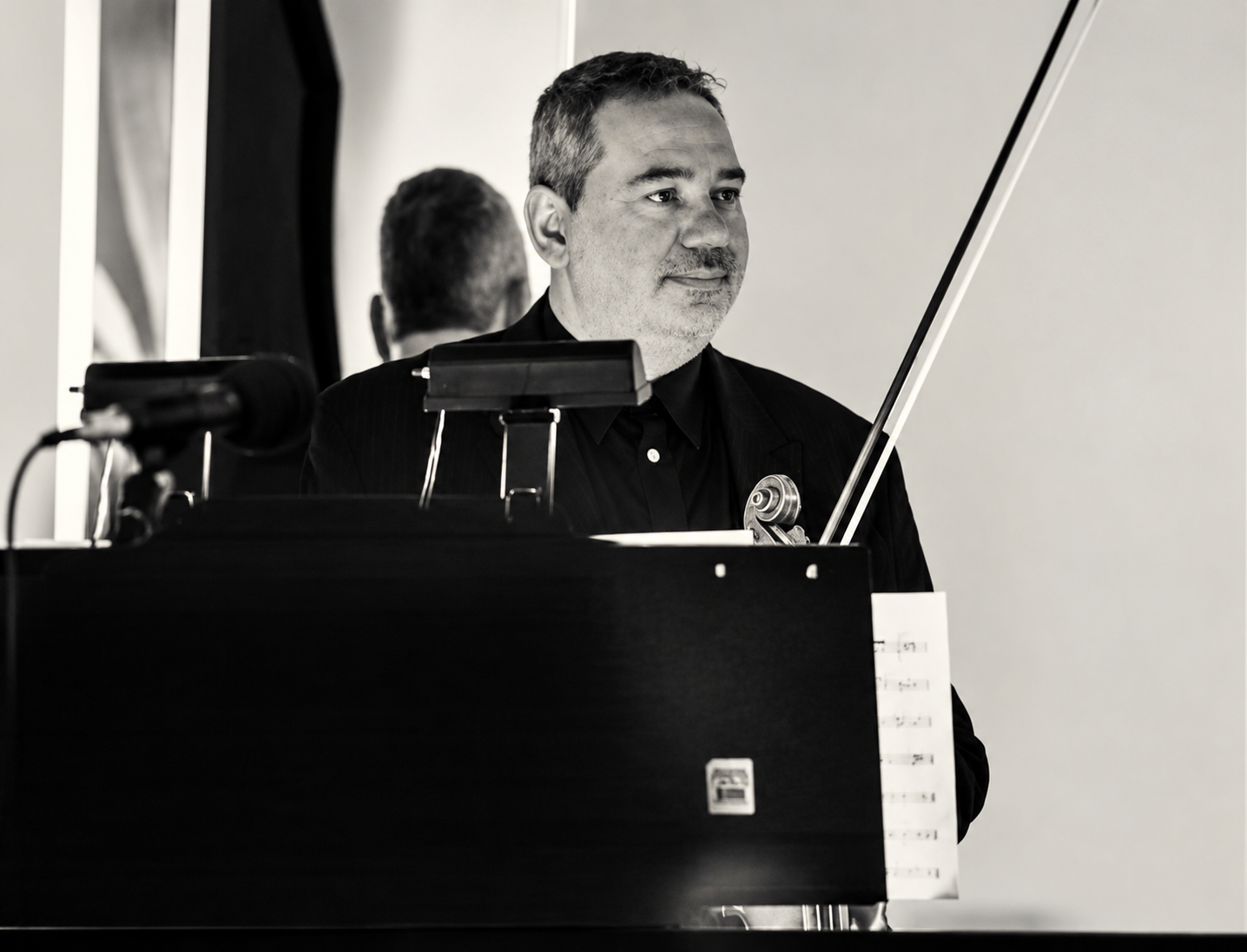 A man in a black suit playing a violin behind a music stand, with a mirror reflecting the back of his head.