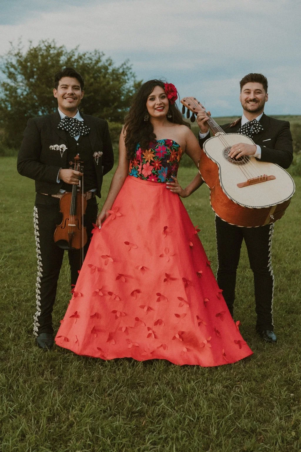 A woman in a vibrant, floral embroidered dress with a full red skirt stands in the center, flanked by two men in traditional Mariachi attire holding a violin and a guitar, standing outdoors on a grassy field with trees in the background.
