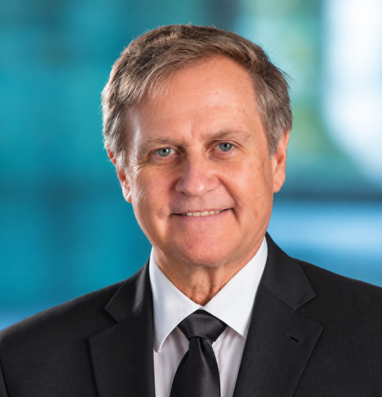 A middle-aged man with light skin, blue eyes, and light brown hair, wearing a black suit, white shirt, and black tie, smiling at the camera against a blurred blue background.