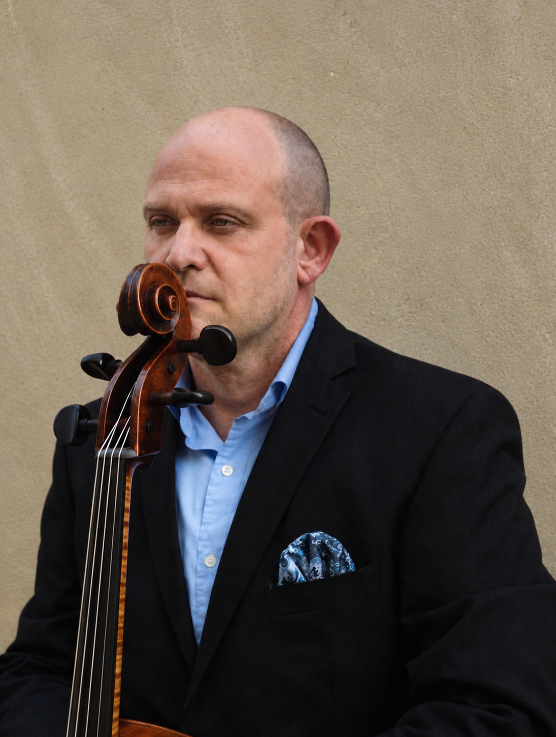 A man in a black suit and light blue shirt with a patterned pocket square, holding a cello, standing against a beige wall.