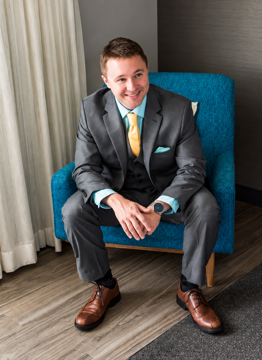 A smiling man in a gray suit, yellow tie, and brown shoes sitting on a blue armchair.