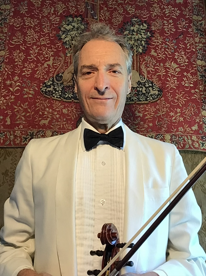 A man dressed in a white tuxedo with a black bow tie, holding a violin and bow, standing in front of a decorative tapestry with floral patterns.