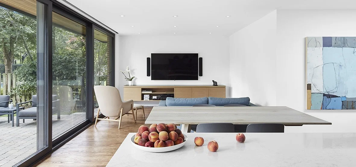 Modern living room with a large sliding glass door, a white armchair, a TV on the wall, a blue couch, a wooden sideboard, a painting, and a bowl of peaches on the kitchen island.