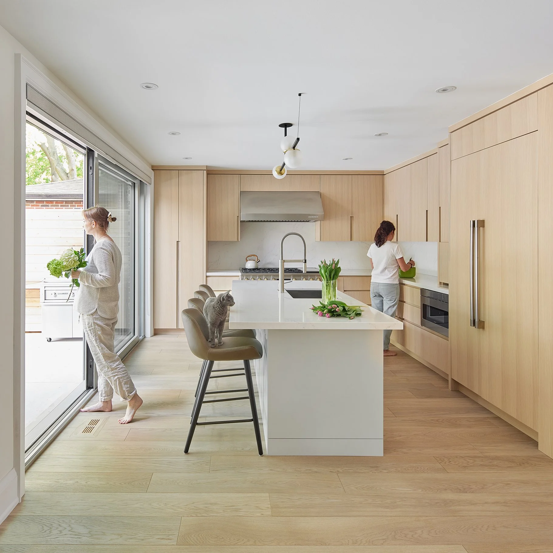 Modern kitchen with light wood cabinets, white island, and two women; one near sliding glass door holding lush green vegetables, another by the counter prepping food; gray cat on bar stool; flowers on island; outdoor patio visible.