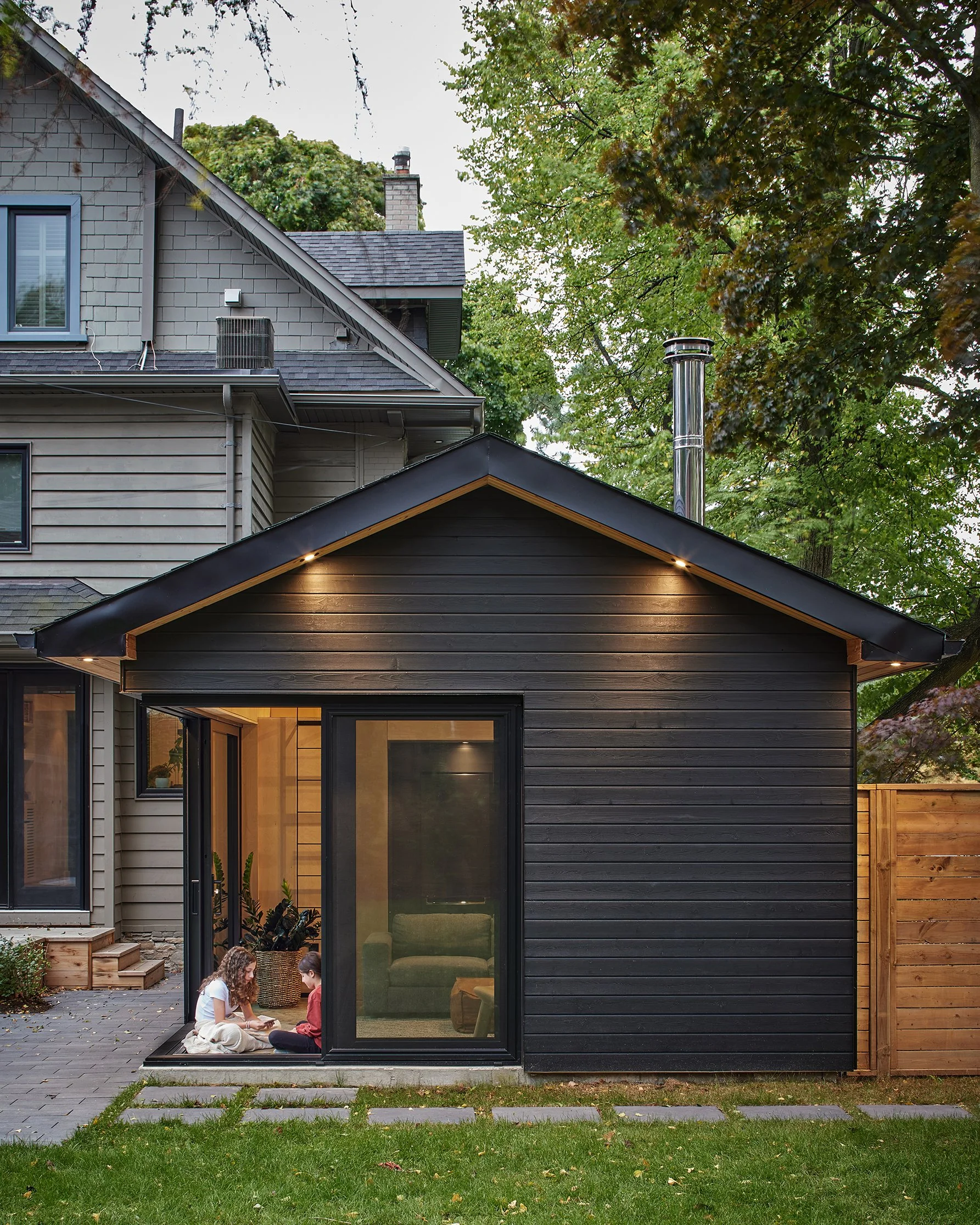 A modern black backyard extension with large glass sliding doors, two children sitting on the doorstep playing a game, green grass in the foreground, and a large tree with green leaves behind. The main house is beige with gray siding, and there is a fenced backyard.
