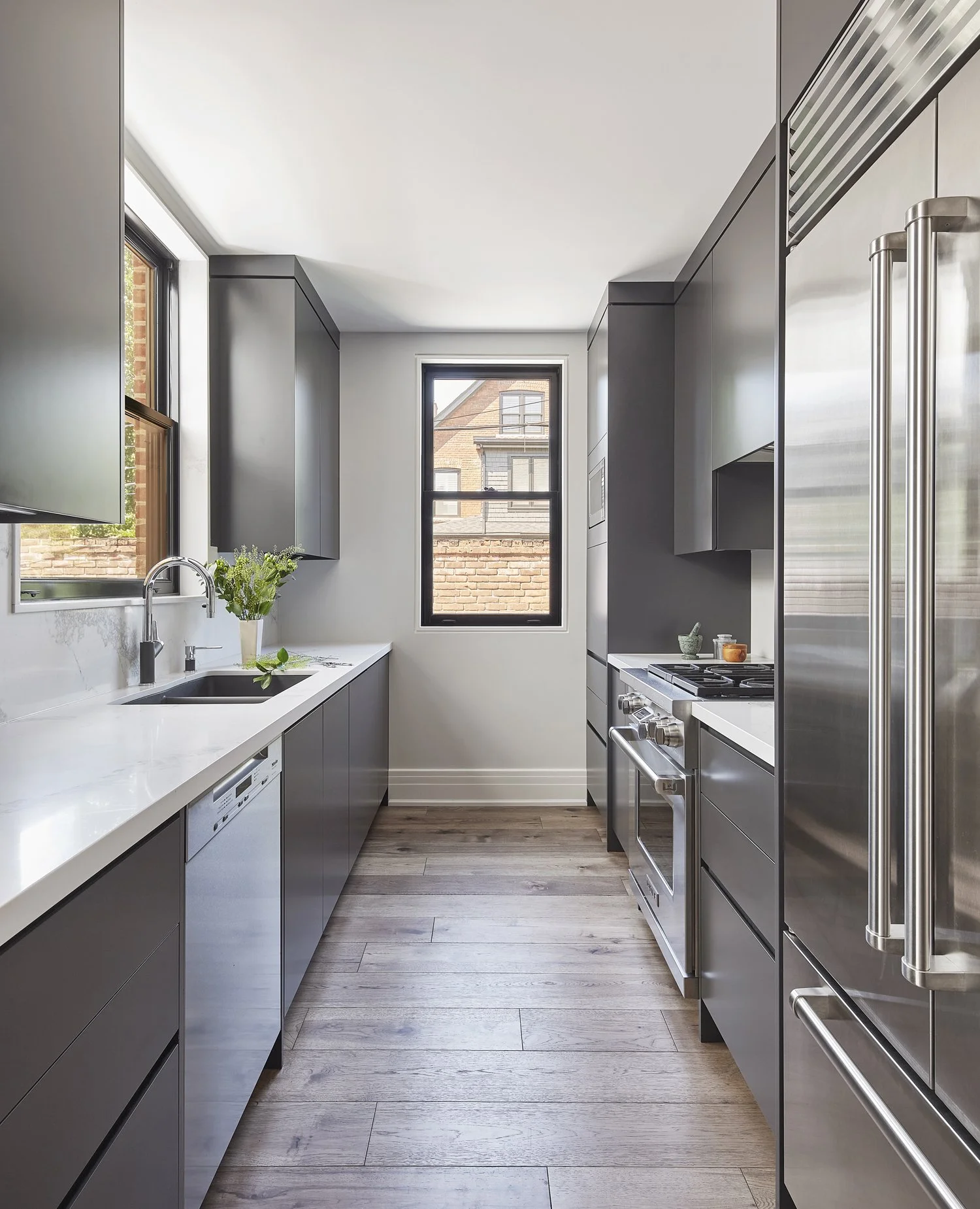 Modern kitchen with white countertops, gray cabinets, stainless steel appliances, and wooden flooring. A window above the sink has a vase of green plants, and another window shows brick buildings outside.