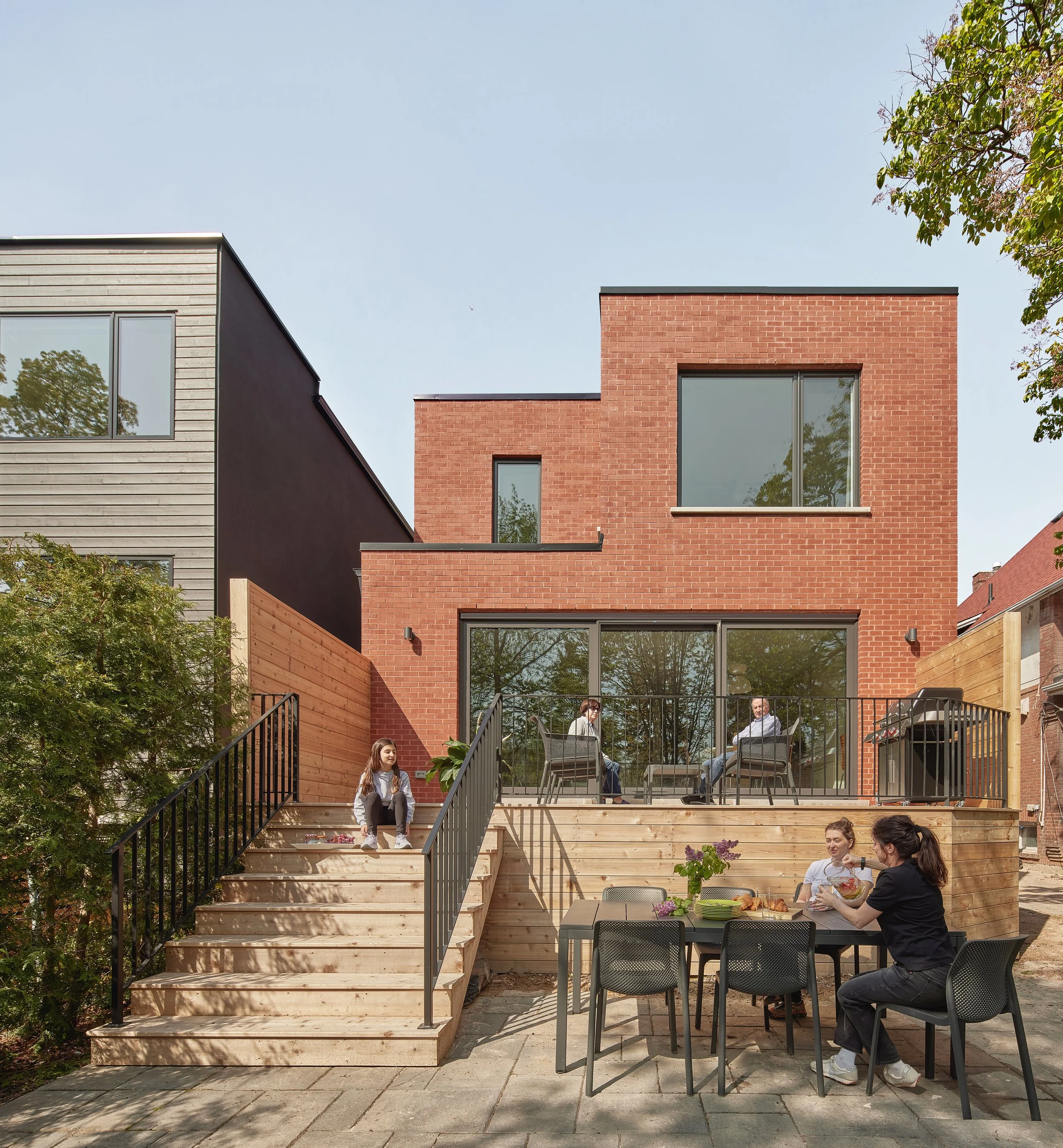 A backyard scene with a modern brick house, outdoor stairs leading to a deck, and a patio table with four women eating, while three people sit on chairs on the deck.