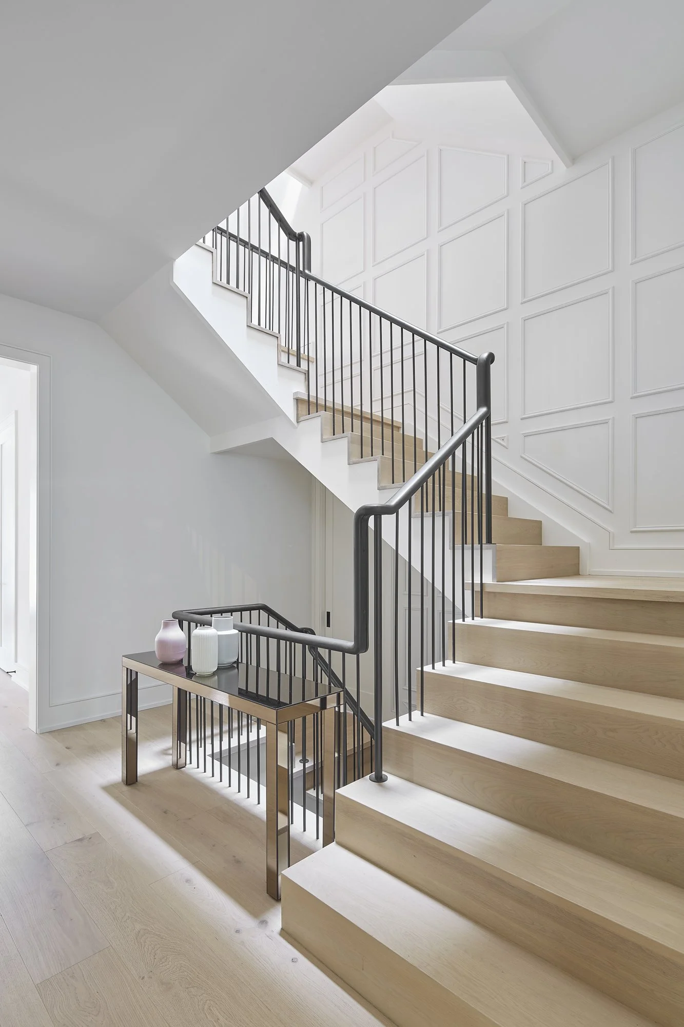 Interior view of a modern staircase with light wood steps and black metal railing, adjacent to a white wall with decorative paneling.
