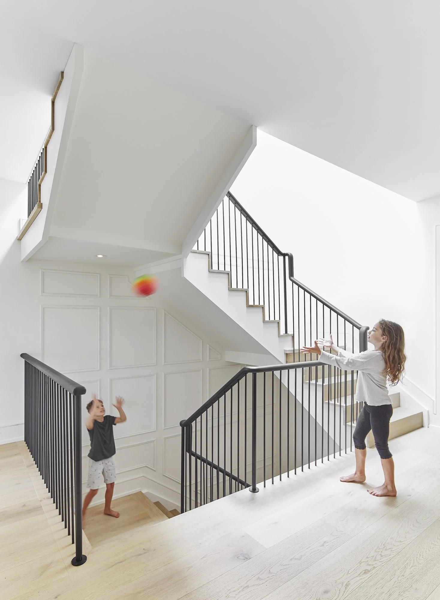 A woman and a young boy playing catch with a small red, green, and yellow ball inside a bright, white, modern home with a staircase and black metal railing.