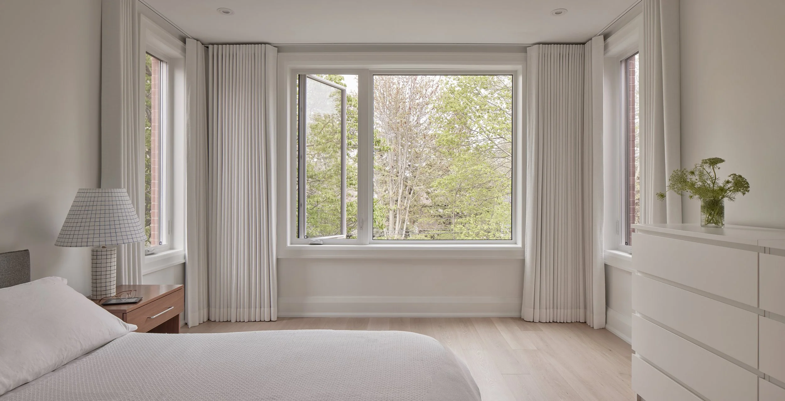 A bright bedroom with white walls, light wood flooring, and large windows with white curtains revealing trees outside. There is a bed with a white quilt, a nightstand with a checkered lampshade, and a white dresser with a glass vase of flowers.