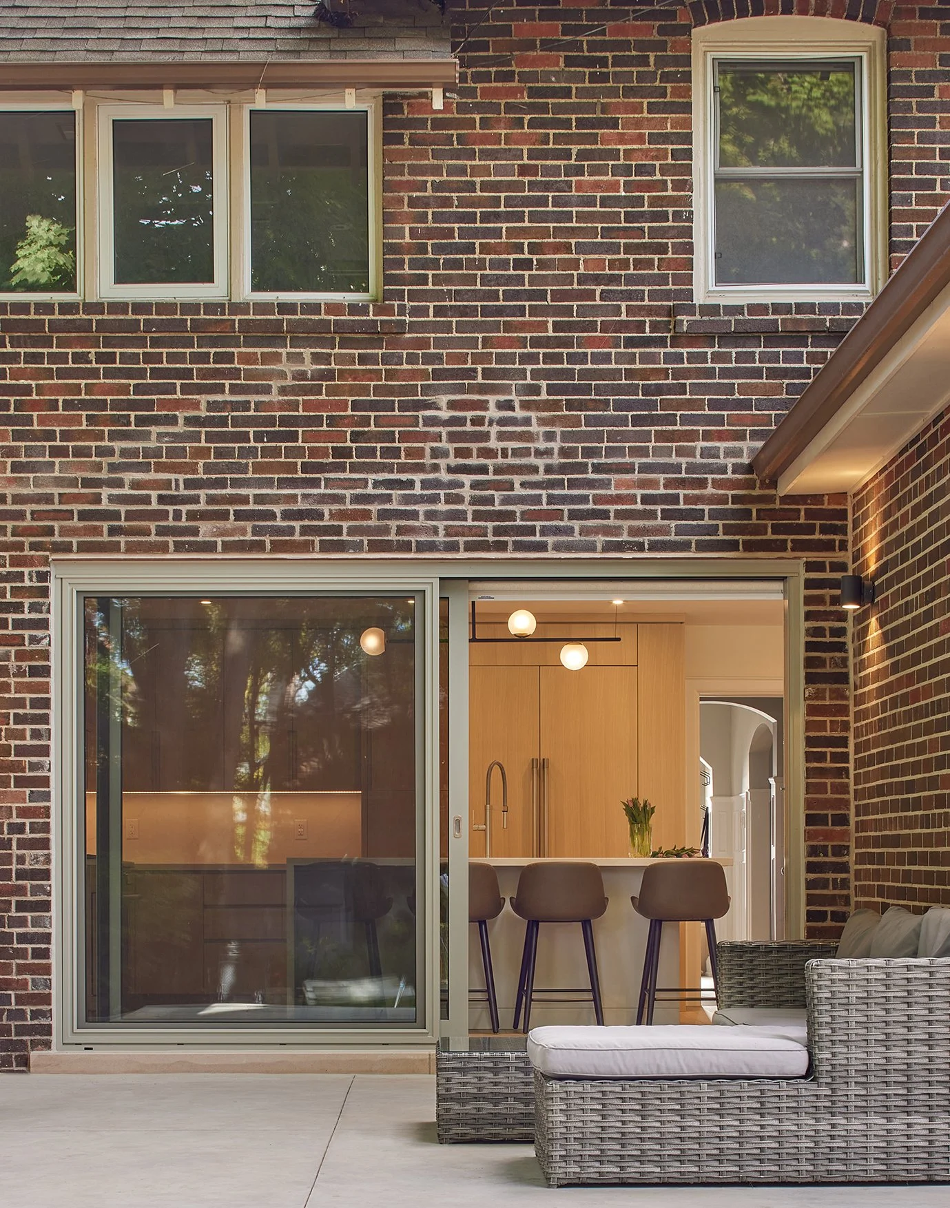 Backyard patio with a brick house, large sliding glass door, outdoor wicker furniture with a cushion, and an open view into a kitchen with wooden cabinets and bar stools.