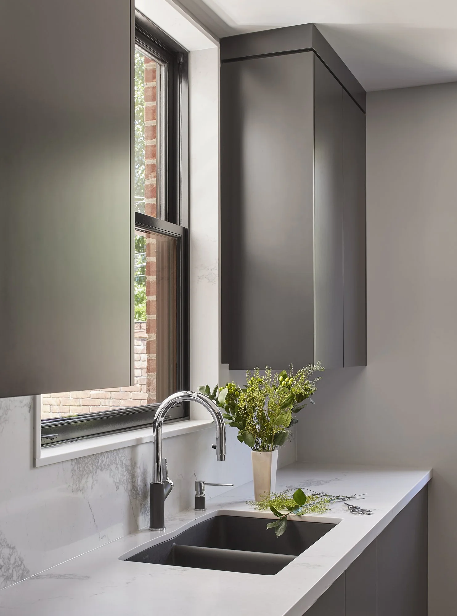 Modern kitchen sink with a silver faucet, white marble countertop, green bouquet in a white vase, located by a window with a brick wall outside.