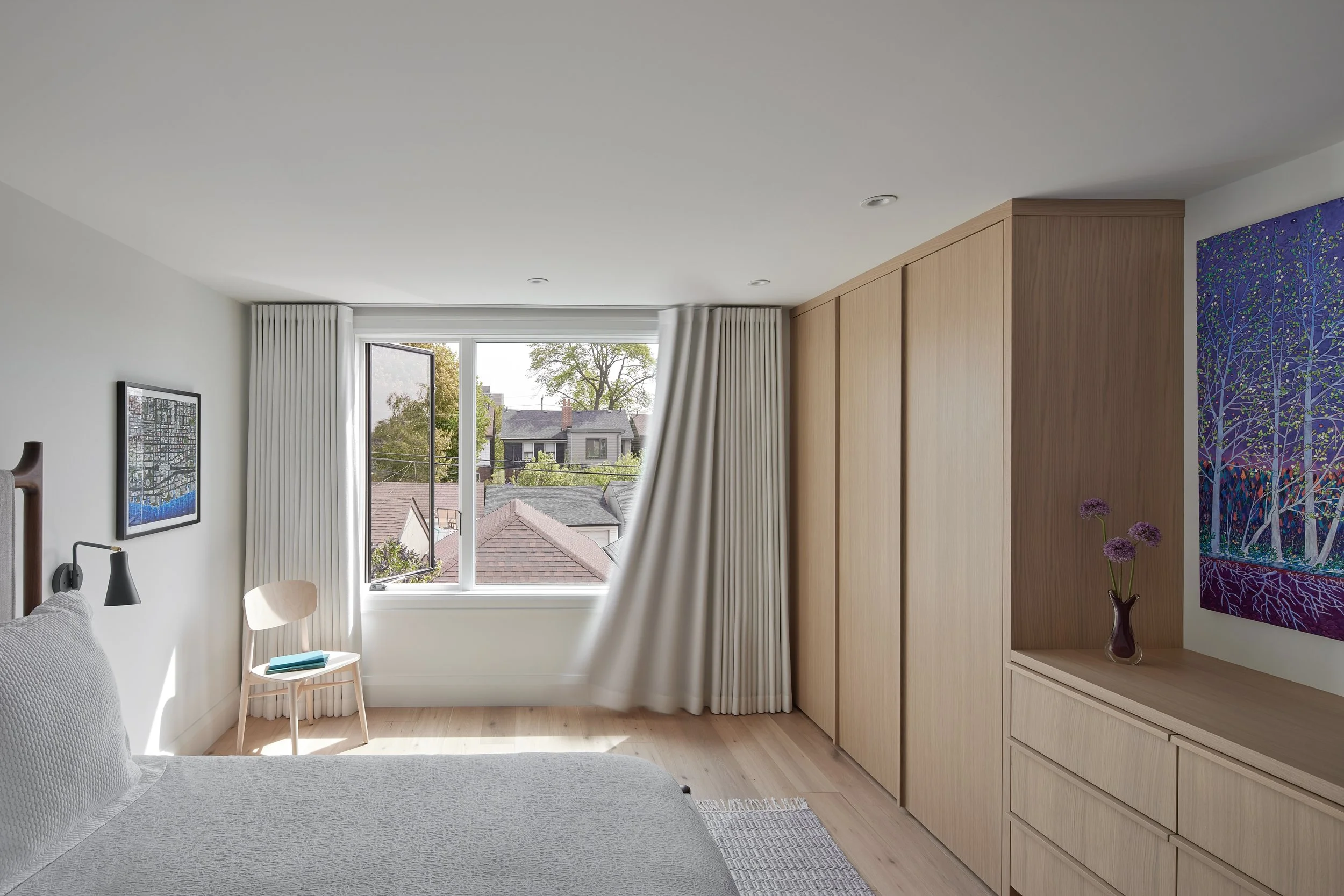Bedroom with large window, white curtains, wooden furniture, a bed with white bedding, a small chair with books on it, colorful wall art, and a vase with purple flowers.