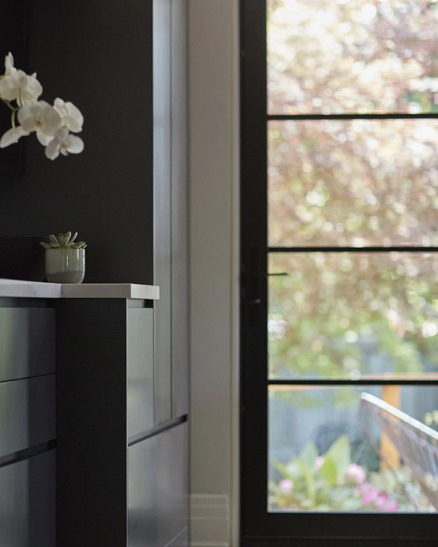 Part of a modern kitchen with dark cabinet drawers, a white countertop with a small potted succulent, and a large glass door showing an outdoor garden with trees and flowers.