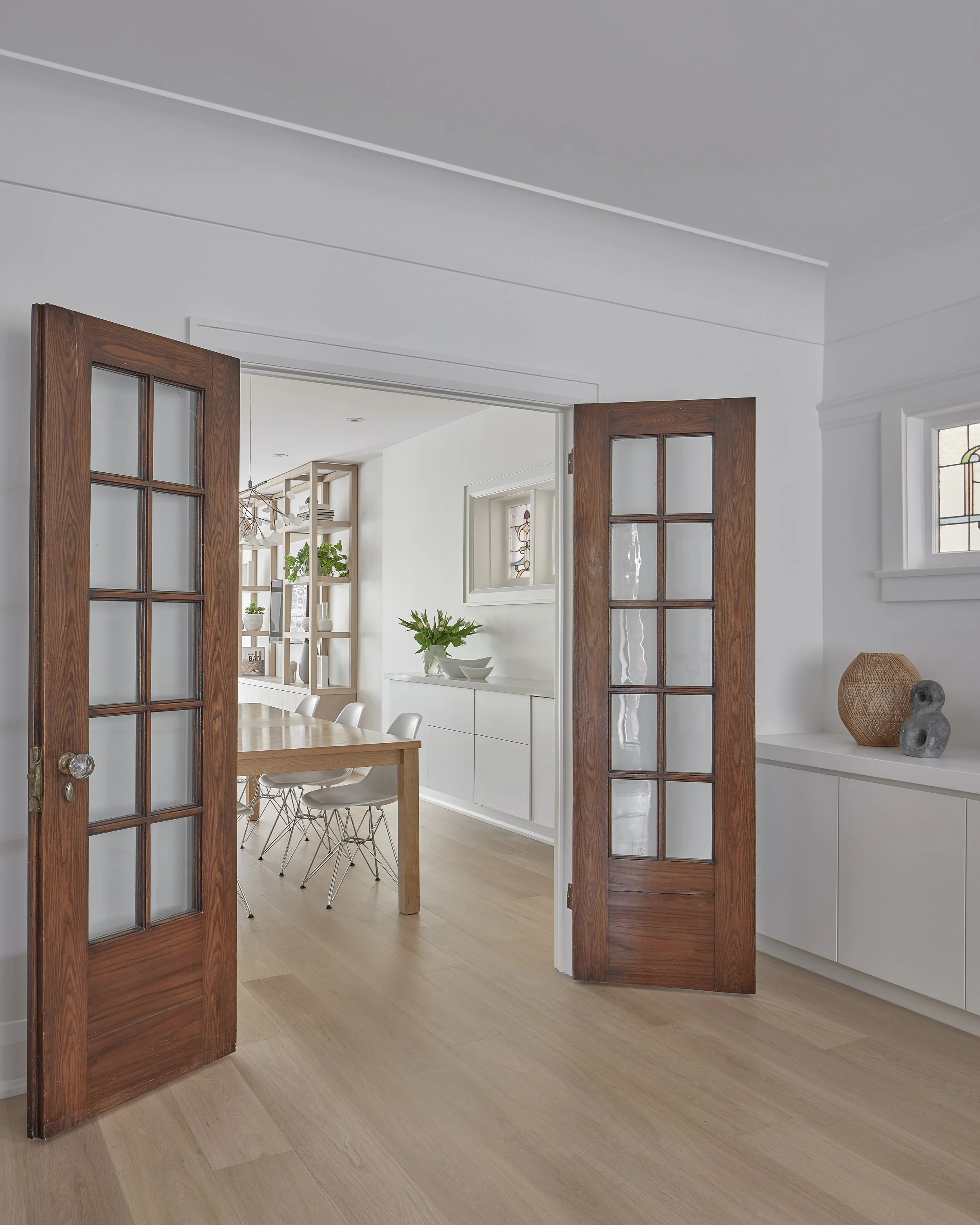 Interior view of a modern dining room with double glass-paneled wooden doors opening to a light, minimalist space with a dining table, chairs, and decorative items.