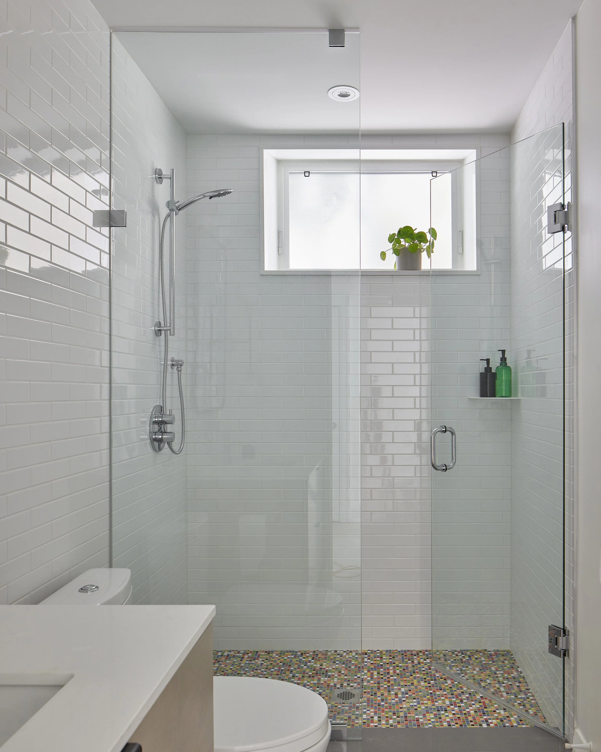 Modern bathroom with a walk-in shower, white tiled walls, a small window with a potted plant, and a colorful mosaic tiled floor.