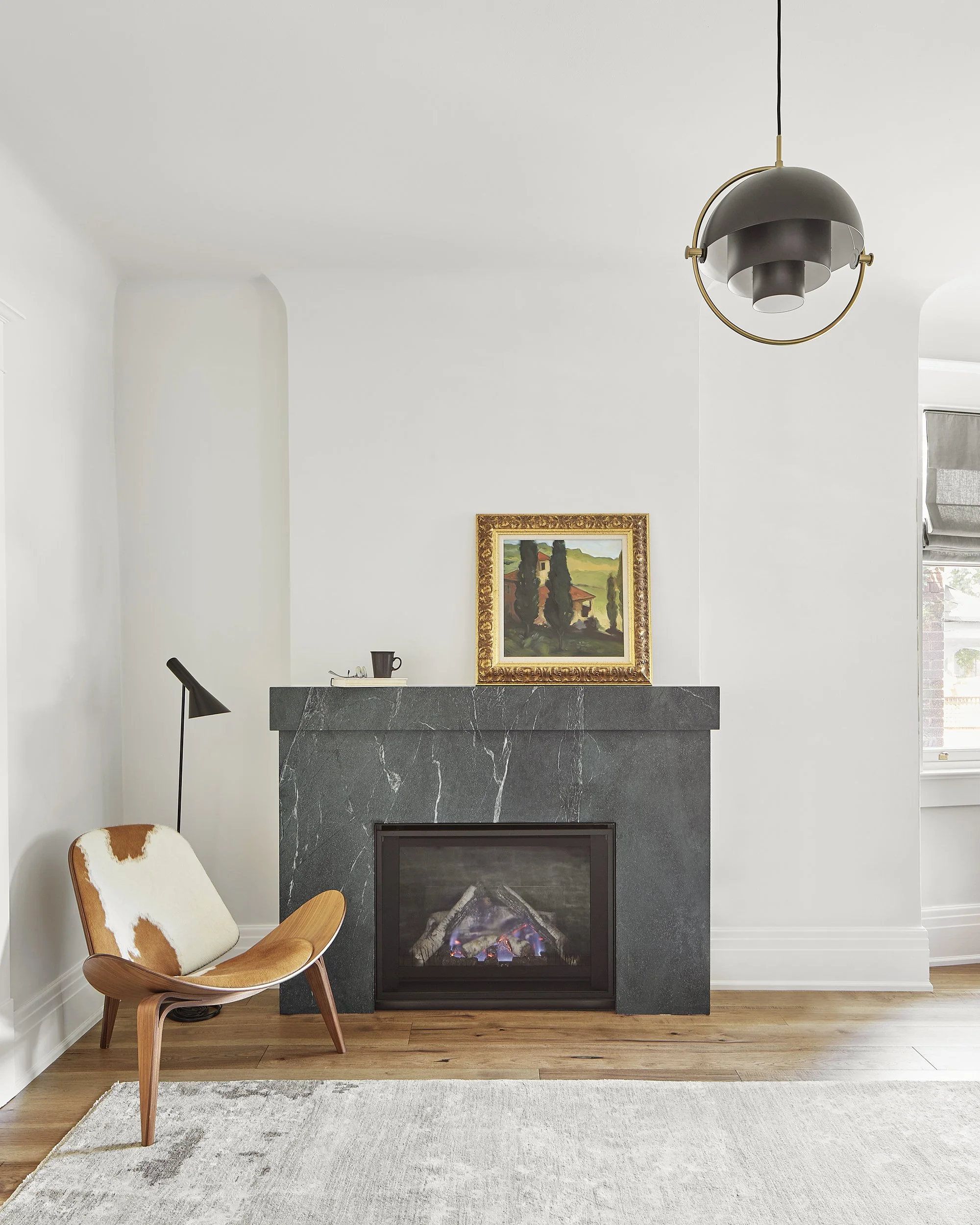 Living room with a black fireplace, a modern chair with cowhide upholstery, and a painting on the mantel, illuminated by a ceiling light fixture and natural light from a window.