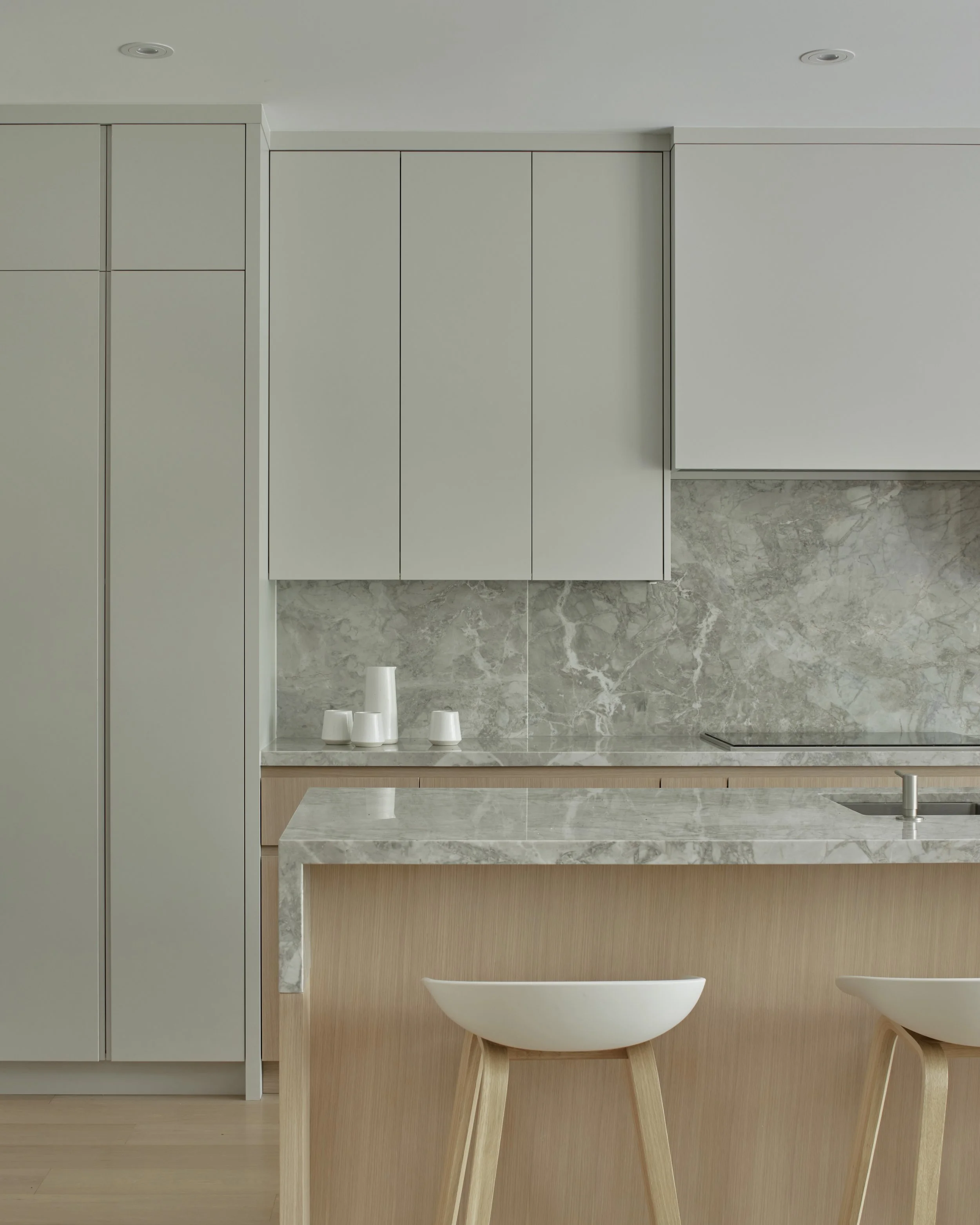 Minimalist kitchen with light gray cabinets, a marble backsplash, and a marble kitchen island with light wood accents. Two white stools with wooden legs are at the island. Three white ceramic cups are on the counter.