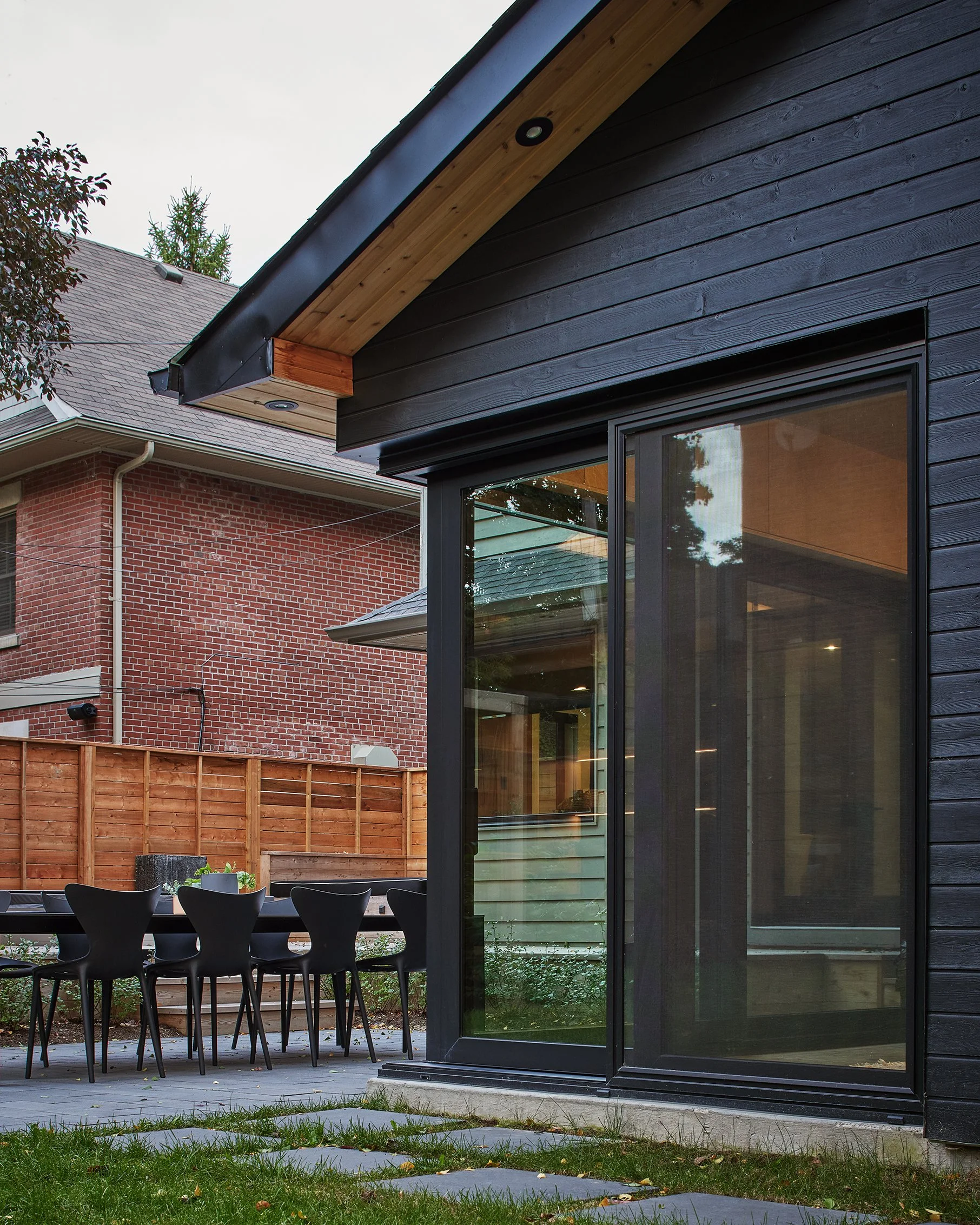 Backyard patio with black dining table and chairs, fenced yard, and modern black house with large glass sliding door.