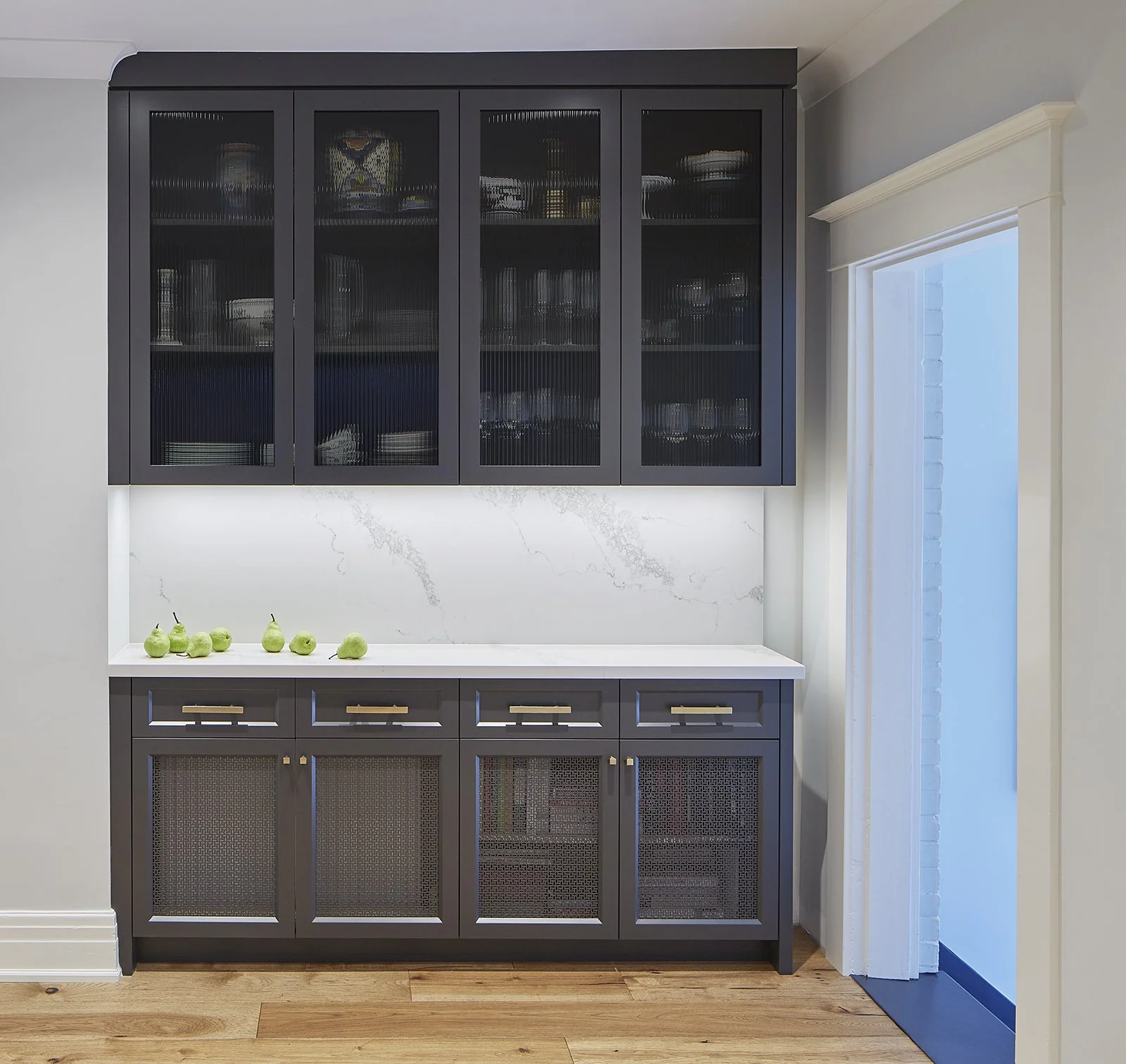 A dark blue kitchen cabinet with glass doors above and solid doors with cane panels below, white marble backsplash, and a white countertop with green pears on it. To the right, there is a white frame around a glass door or window, and a wooden floor.