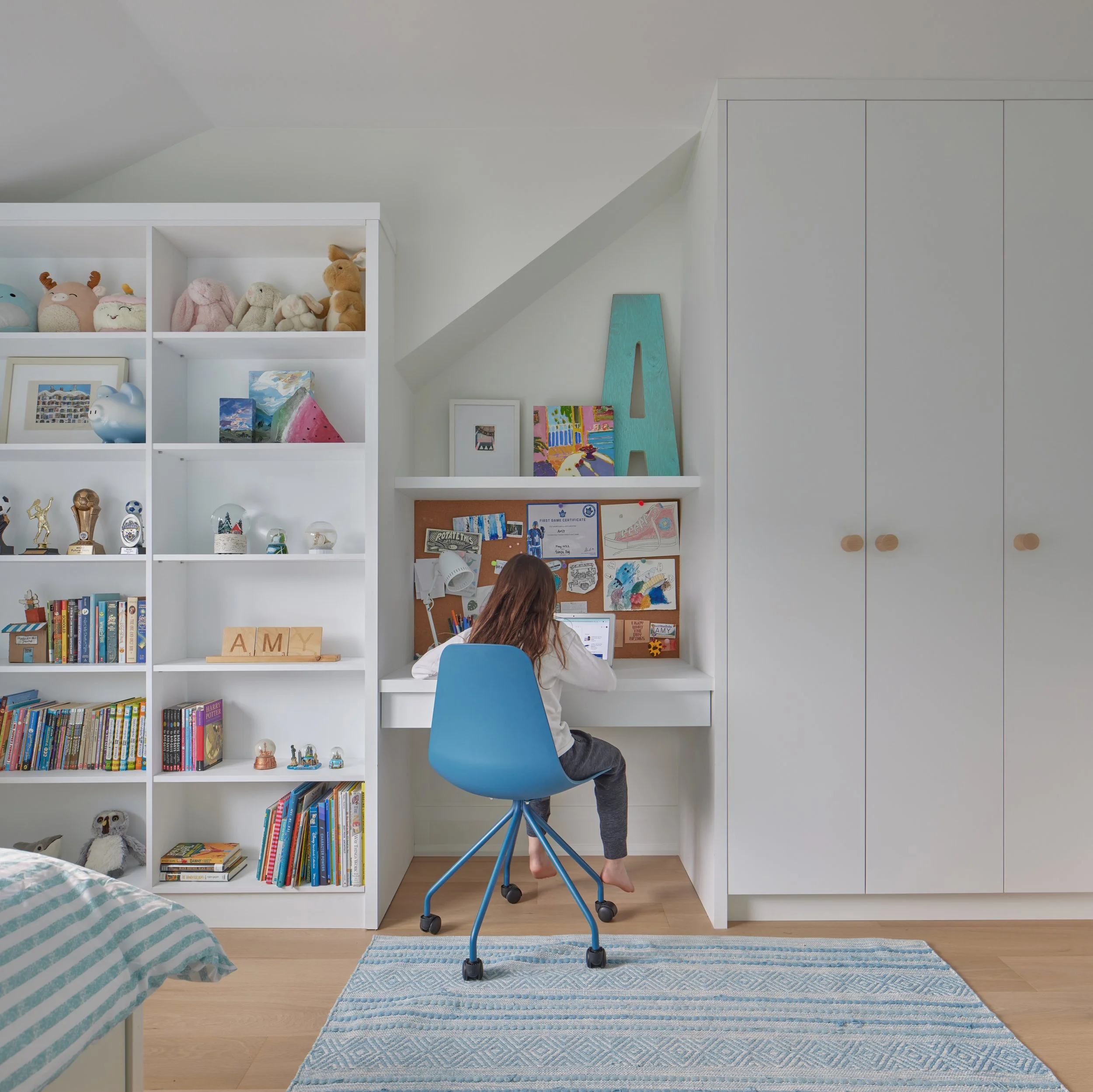 Child sitting at a white desk, working on a computer, in a bedroom with white bookshelves filled with toys, books, and decorations, and a blue chair, with a blue patterned rug and a bed with striped bedding in the foreground.