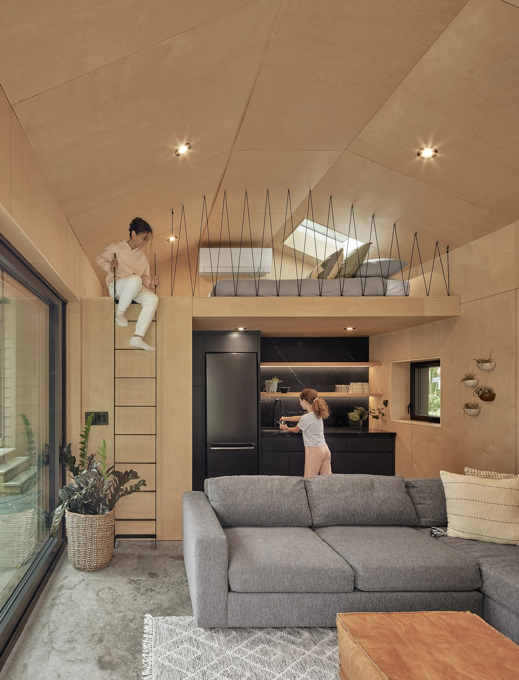 Interior of a modern living space with a living room, kitchen, and a lofted sleeping area. Two children are playing; a girl is on a ladder leading to the loft, which has a bed with pillows and a skylight. A woman is in the kitchen near the sink, and there are plants and wall decorations visible.
