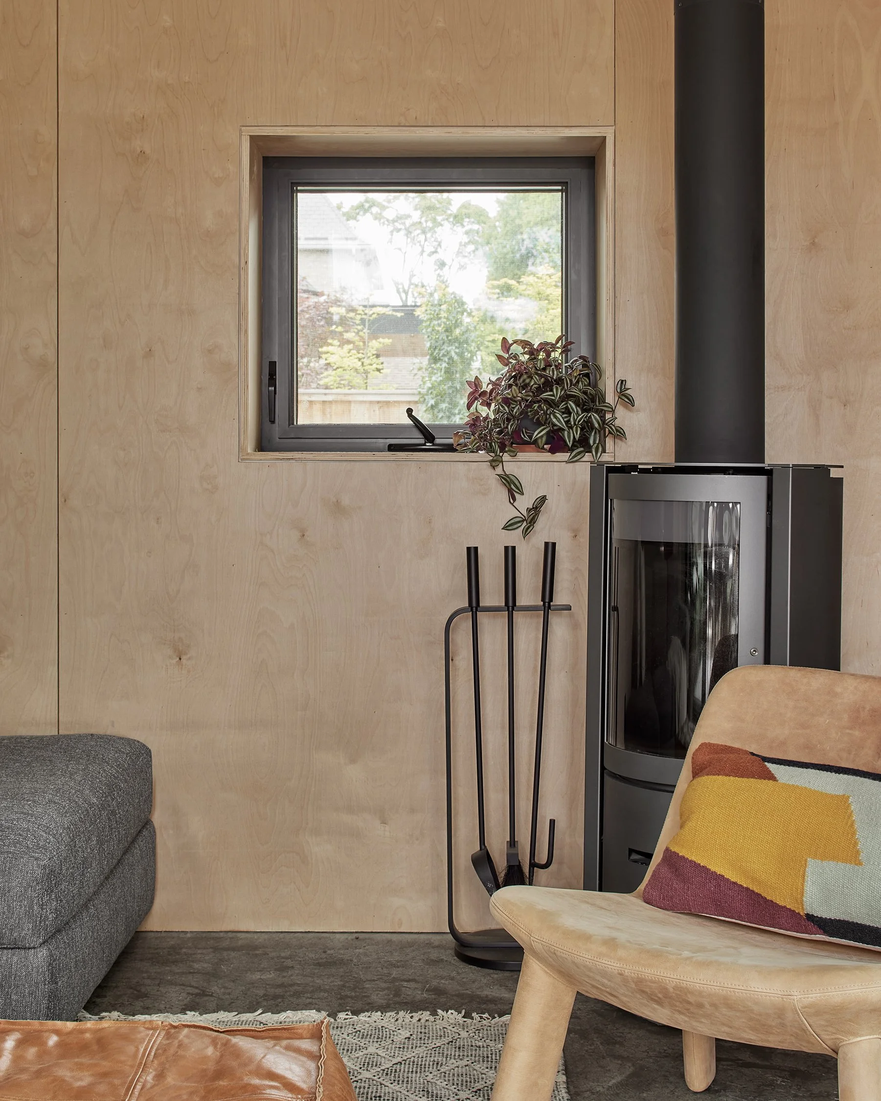 A cozy living room corner featuring a small window above a potted plant, a modern black fireplace with a chimney, a fire toolset, a beige chair with colorful pillow, and part of a gray sofa.