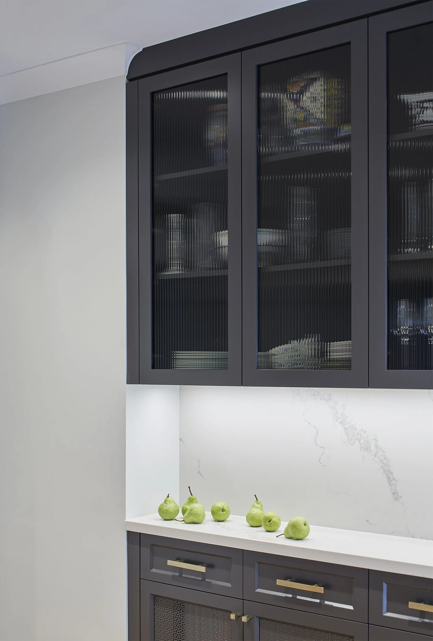 Black kitchen cabinet with glass doors above a white marble countertop with six green pears arranged on it.