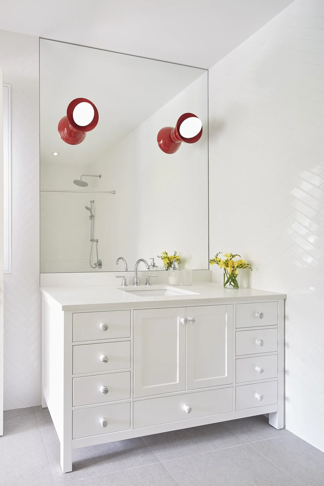 A white bathroom vanity with drawers and a cabinet, a countertop with a sink, and a large mirror above it. Two red wall sconces are reflected in the mirror. Decorative yellow flowers are placed on the countertop.