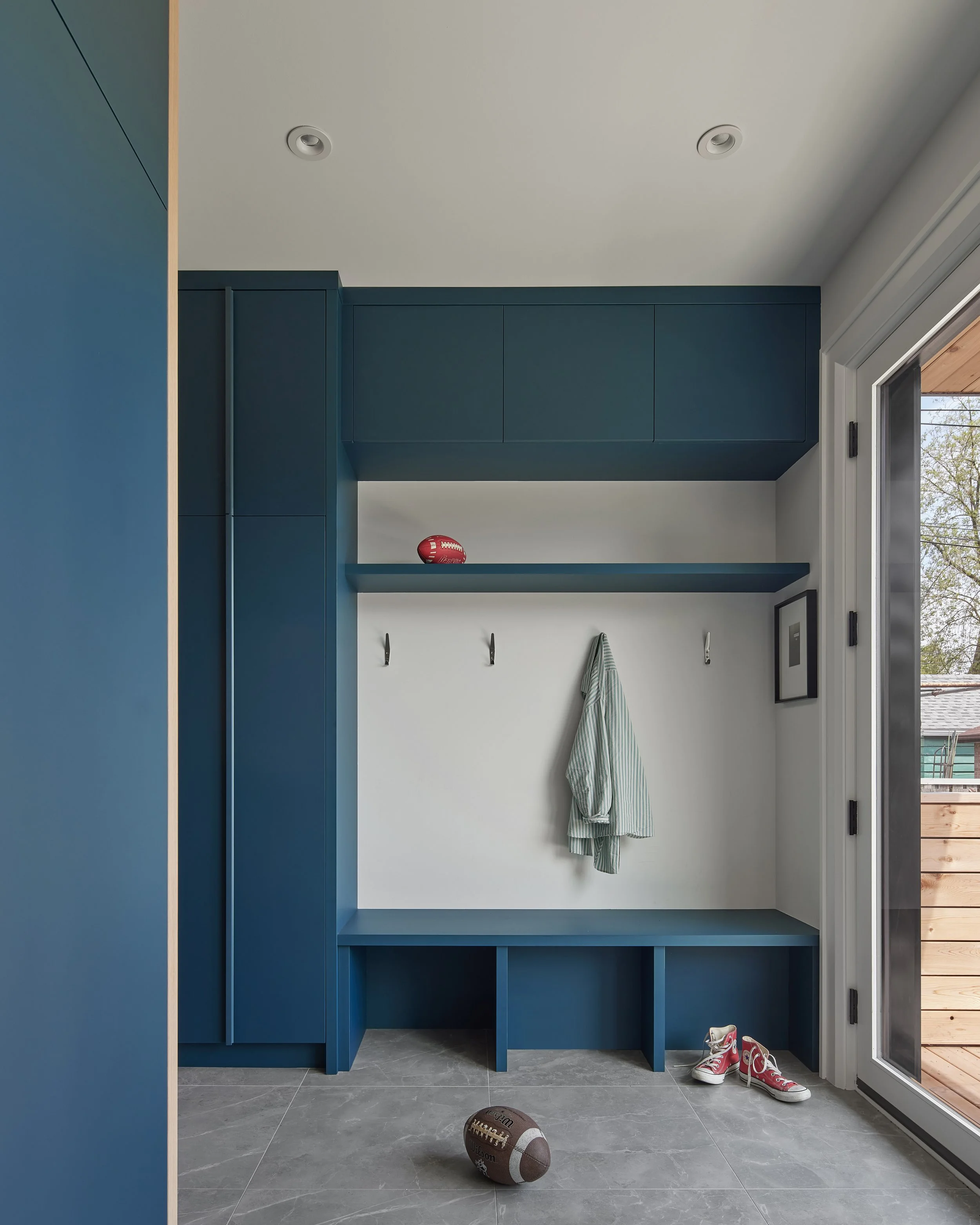 Mudroom with blue built-in cabinets, hooks with a hanging shirt, a football on the floor, and red sneakers near a sliding glass door.