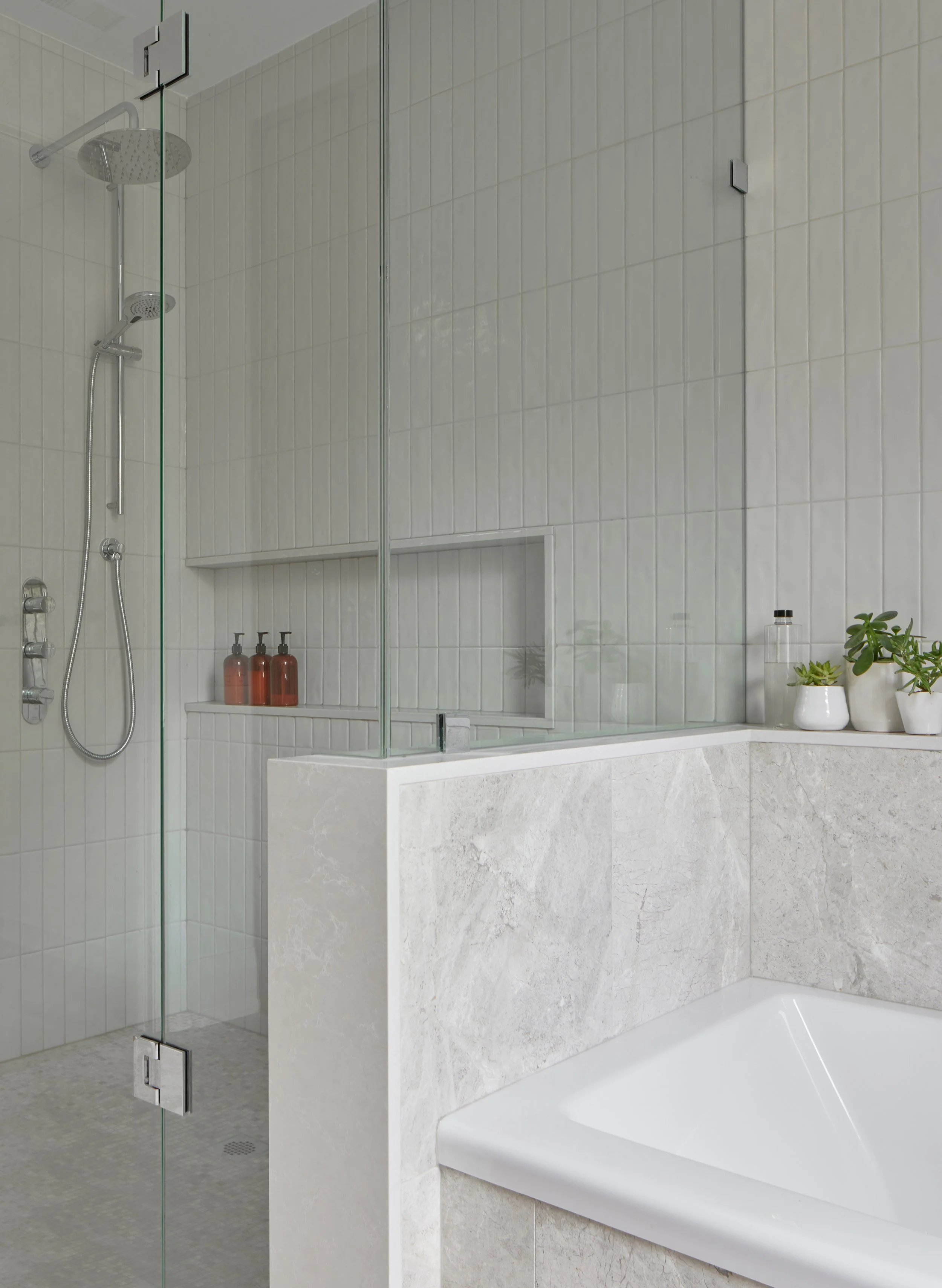 Modern bathroom with a glass shower enclosure and a bathtub. The shower has a rain shower head and a handheld shower. There are three amber-colored bottles on a built-in shelf in the shower area. The bathtub is made of white material with a textured gray panel on the side. The wall behind the bathtub has white tiles, and there are small potted plants and a water bottle on the ledge near the bathtub.