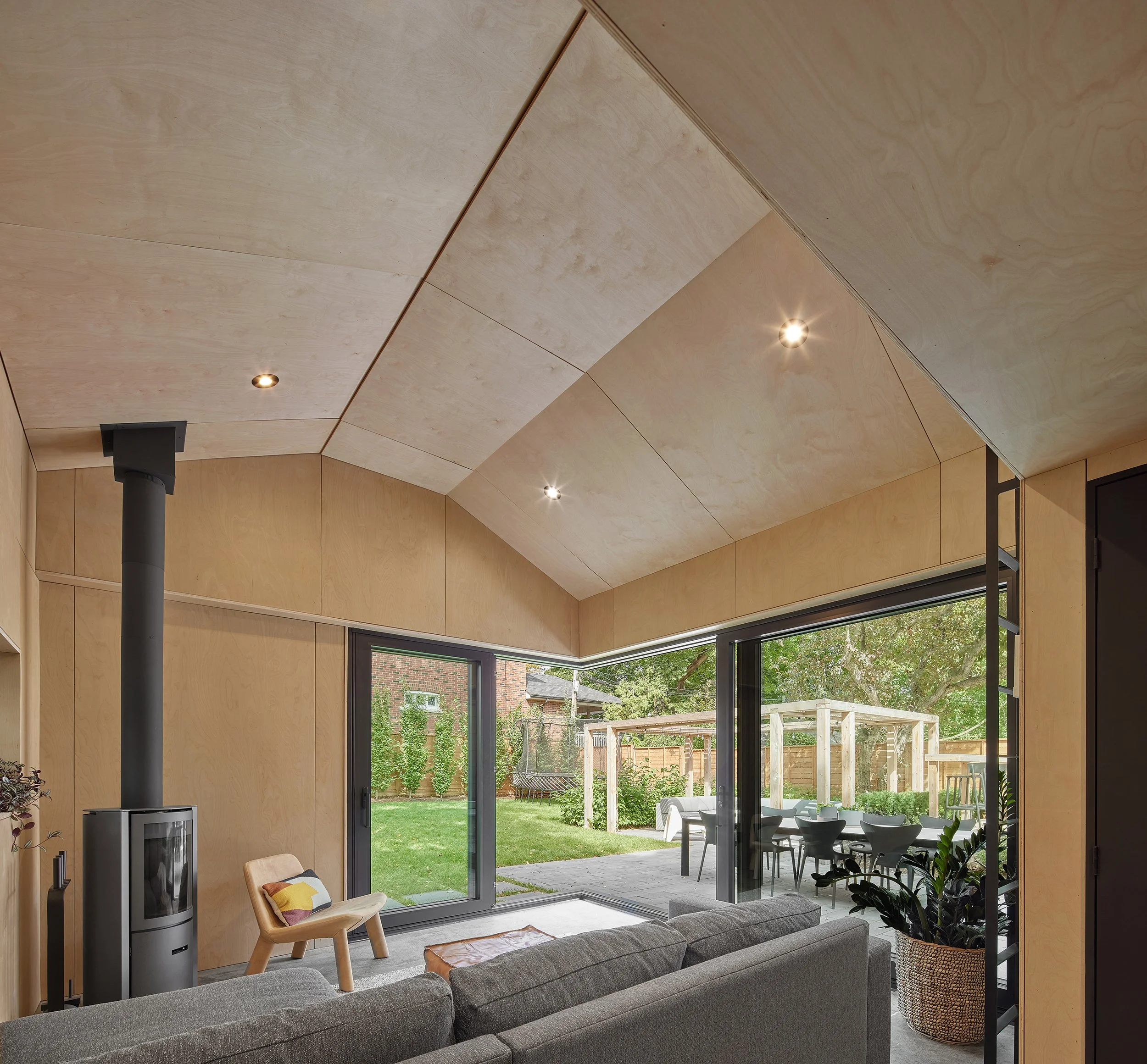 Living room with large sliding glass doors opening to a backyard patio with outdoor dining and garden, wood-paneled ceiling, and modern furniture.