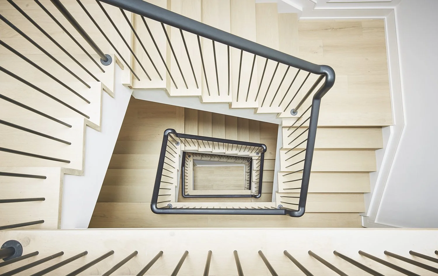 Top-down view of a spiral staircase with wooden steps, black metal handrails, and white walls.