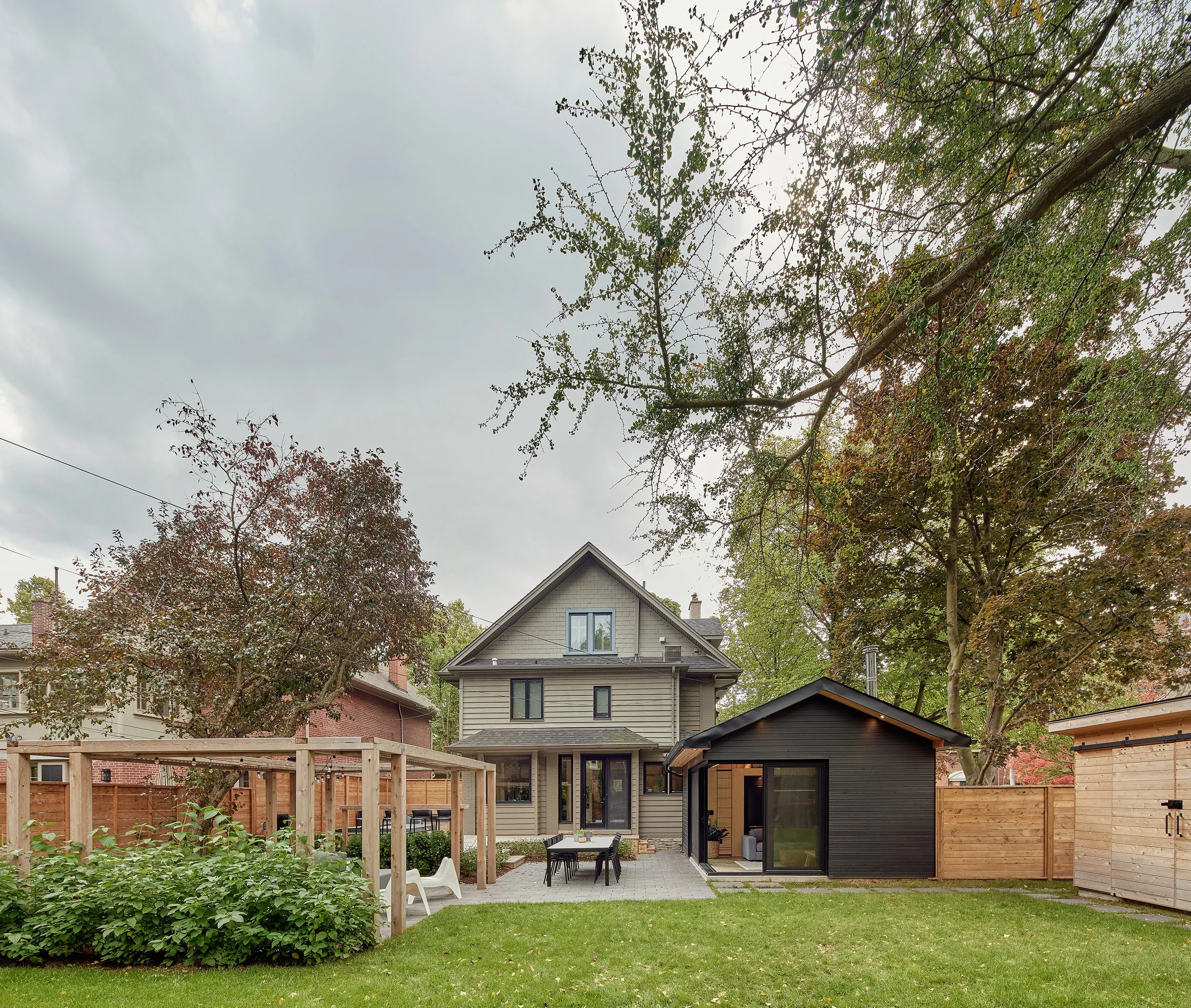 Backyard with a gray two-story house, a small black shed, a wooden pergola, and a landscaped garden with green grass and trees with autumn leaves under an overcast sky.
