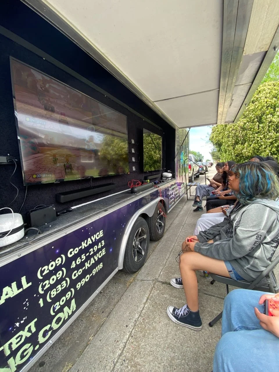 A group of people, mostly teenagers, sitting outdoors on chairs watching a movie or video on a large flat screen television mounted on a mobile trailer. The trailer has contact information and a website, and is parked on a sidewalk near trees and par