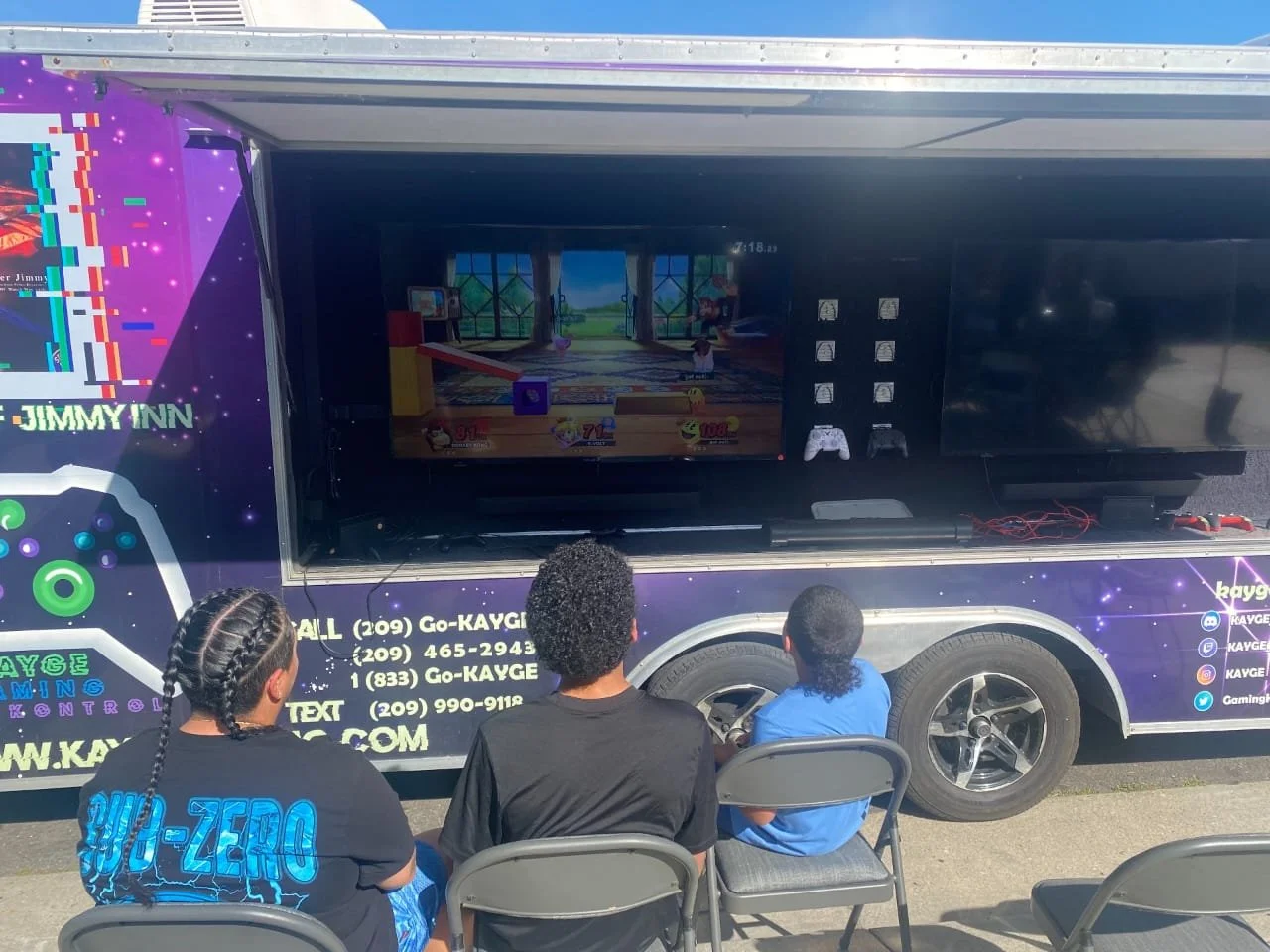 Three children sitting on chairs and playing video games on a mobile gaming truck with large screens outside.