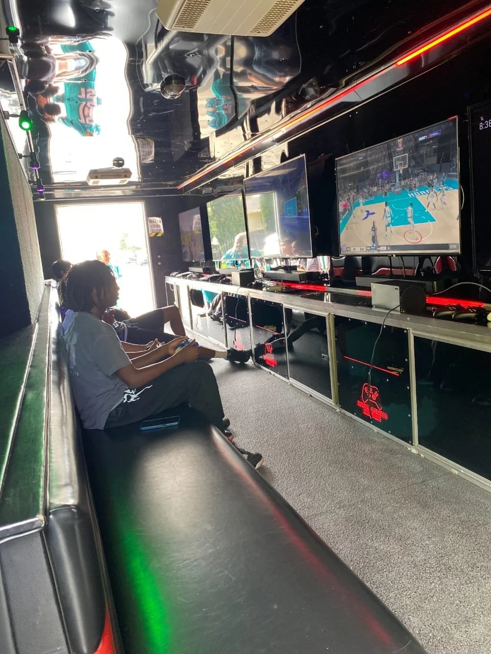 Two children sitting on a black bench playing video games on a console inside a gaming lounge. Multiple large screens display a basketball video game, with a person visible outside through the front door.