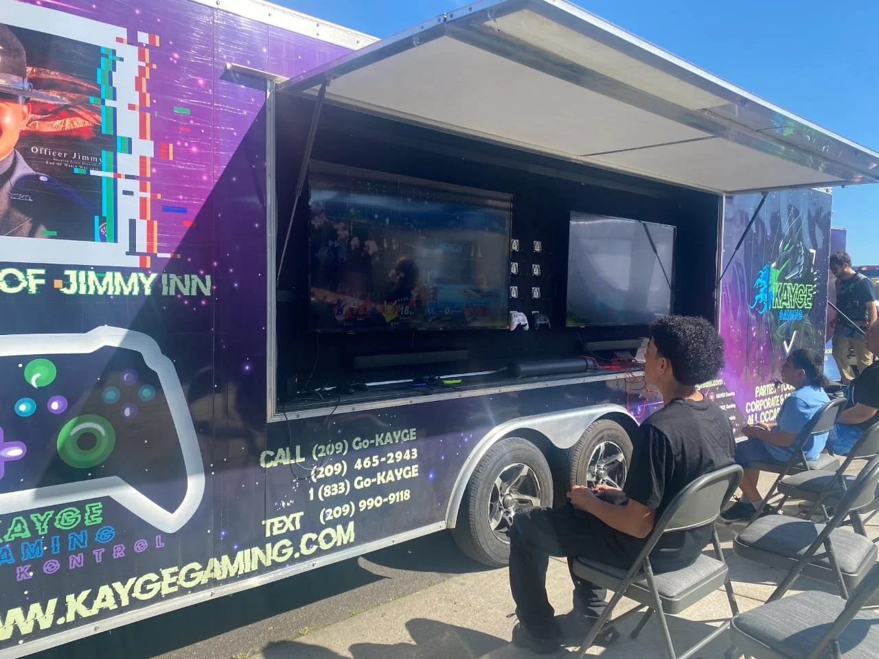 People sitting outside a gaming trailer with screens, controllers, and promotional graphics for Kayge Gaming Control at an outdoor event.