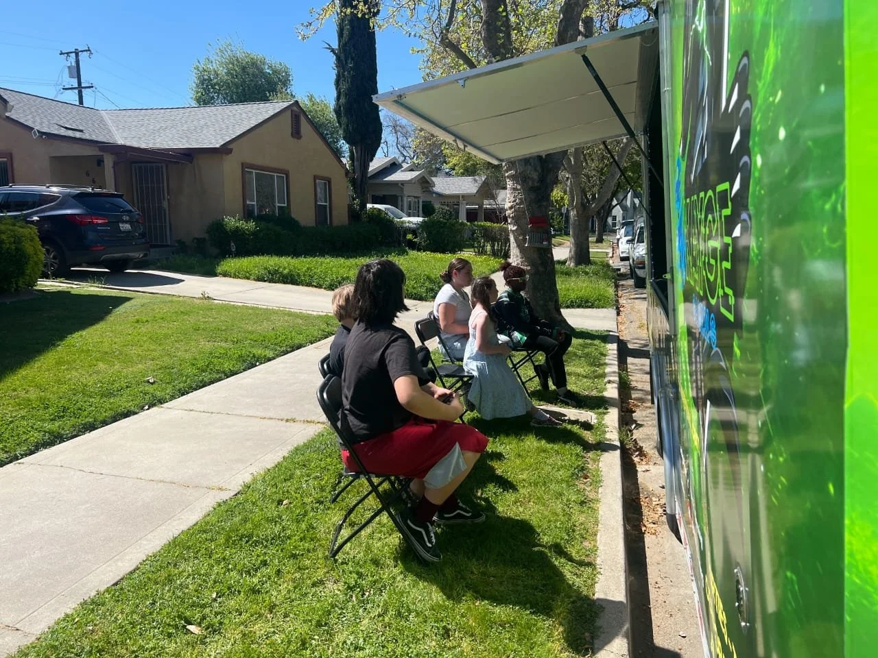 People sitting on chairs outdoors on a sidewalk near a green food truck in a residential neighborhood on a sunny day.