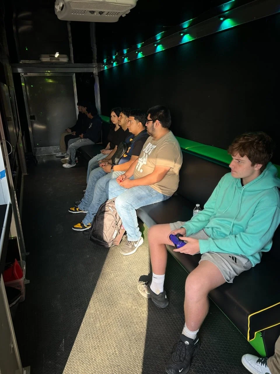 A group of young people sitting on a black bench in a dark room, some holding game controllers, with green lighting along the ceiling.