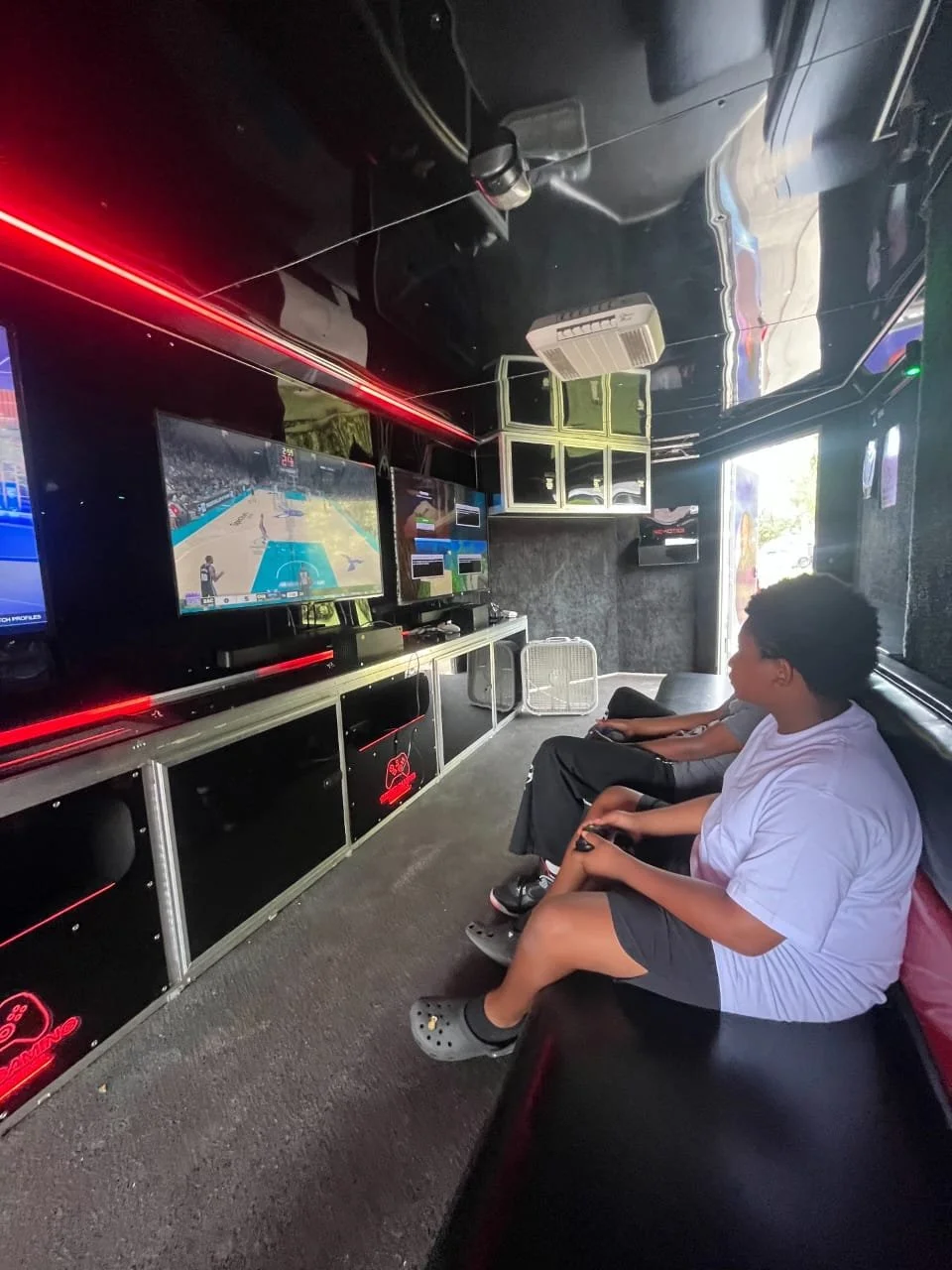 Two boys sitting on a black couch playing video games in an entertainment room with multiple monitors showing a basketball video game.
