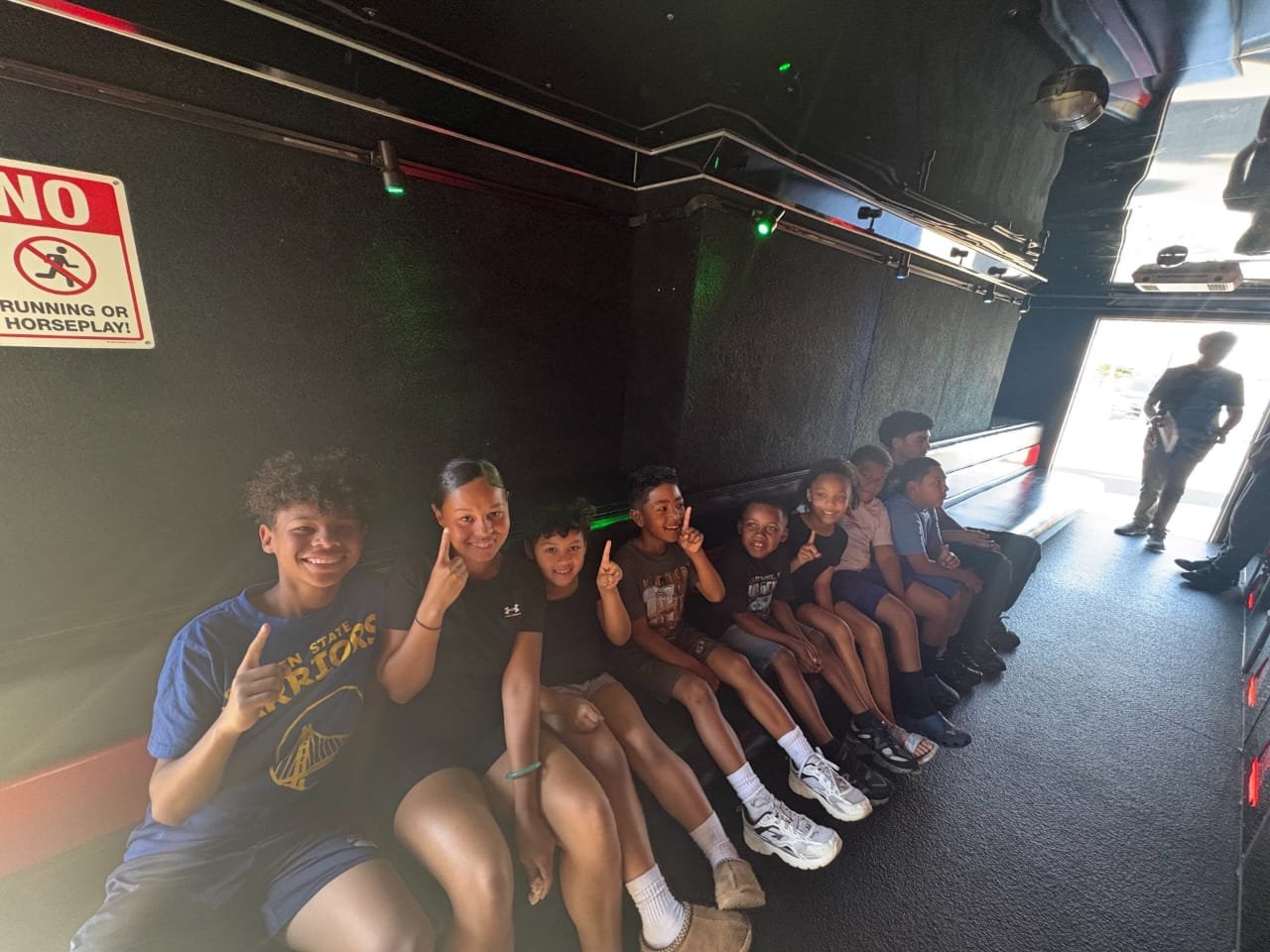 Group of children sitting on a bench inside a facility, smiling and waving at the camera.