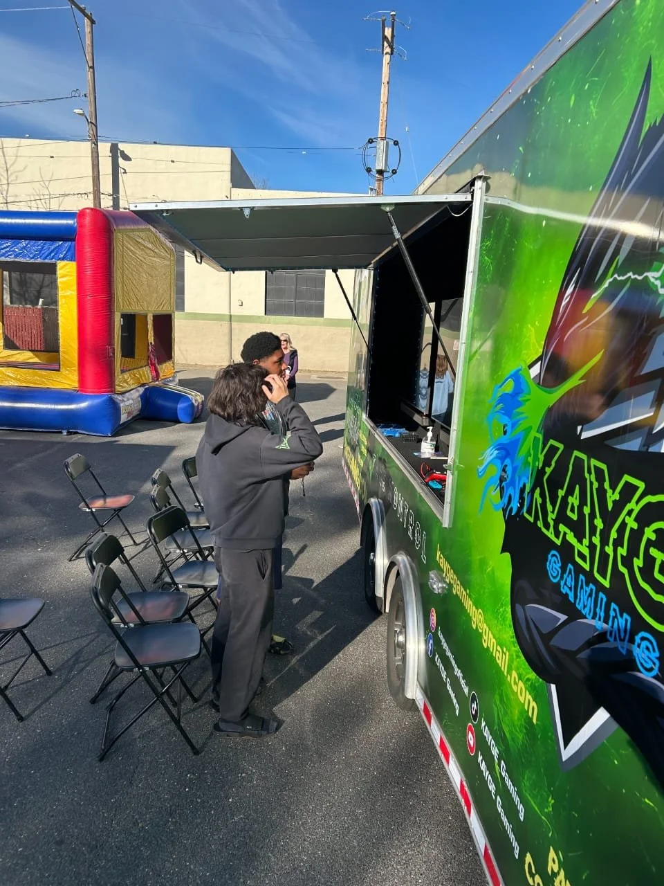 Two people are standing in front of a green gaming truck with a window open, possibly ordering or waiting. Behind them, there is an inflatable bounce house, and a woman is walking in the background. The scene takes place outdoors on a clear day.