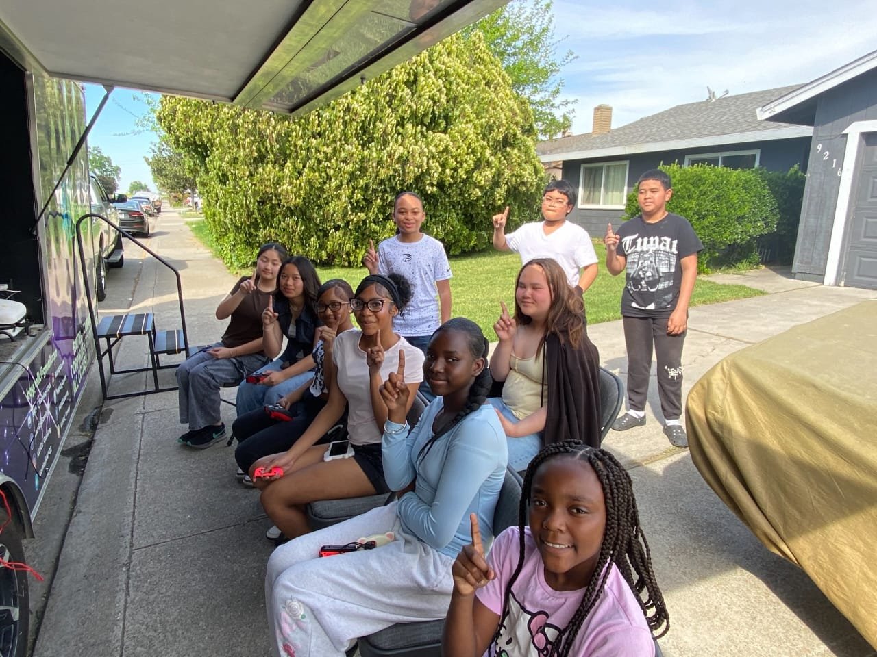 A group of children and teenagers gathered outdoors in front of a building and trees, some sitting and some standing, with many holding up one finger and making gestures.