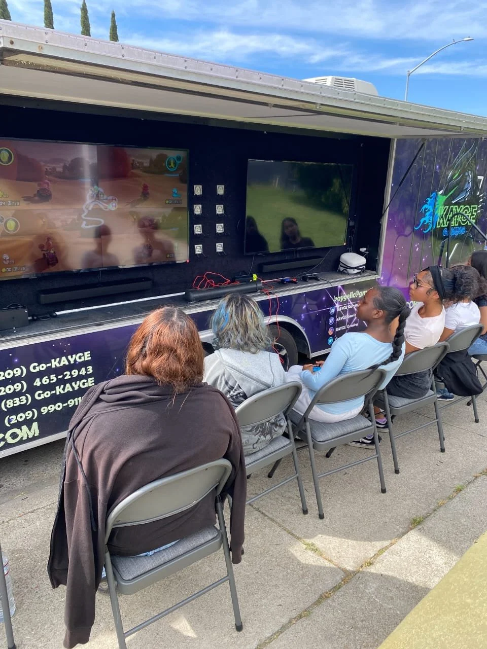 A group of people, including children and adults, sitting on chairs outside, watching a gaming event on two large screens set up on a mobile gaming truck. The truck has a colorful, galaxy-themed design.