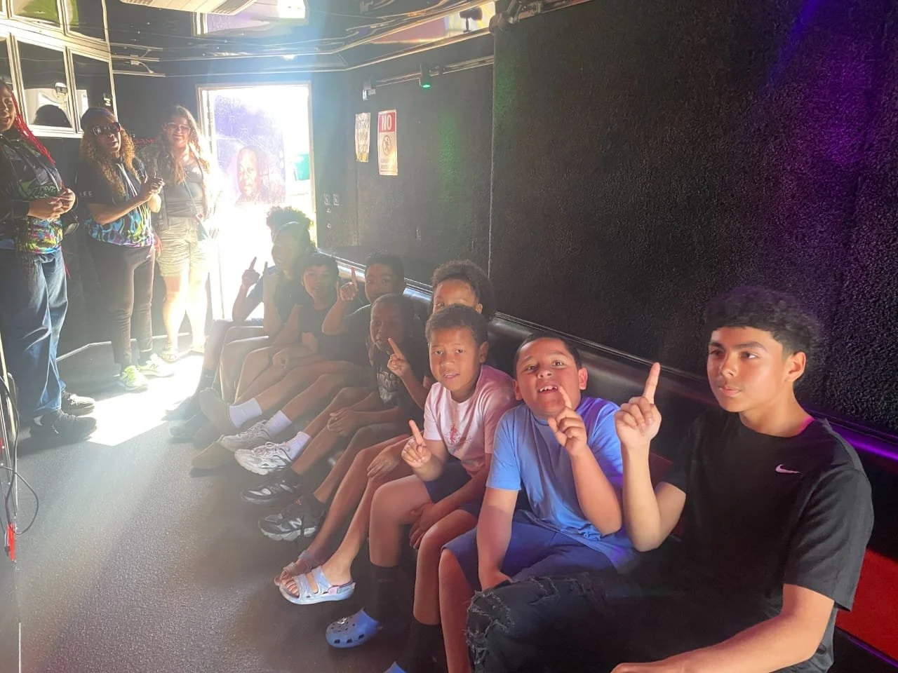 A group of children sitting on a bench inside a room, with some of them making peace signs and looking at the camera. There are people standing near the door, and sunlight coming through the door window.