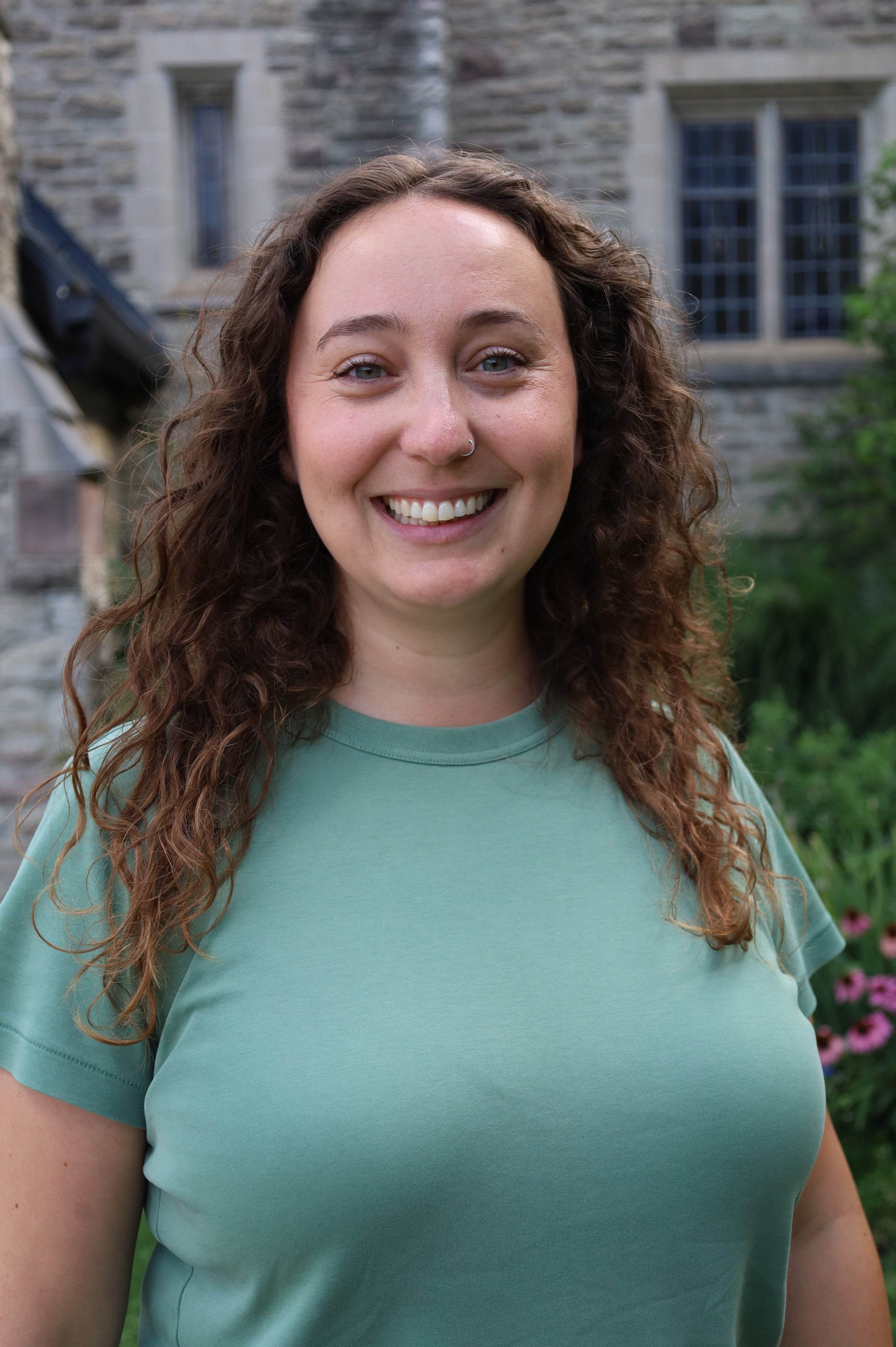 A woman with curly brown hair smiling in front of a brick wall.