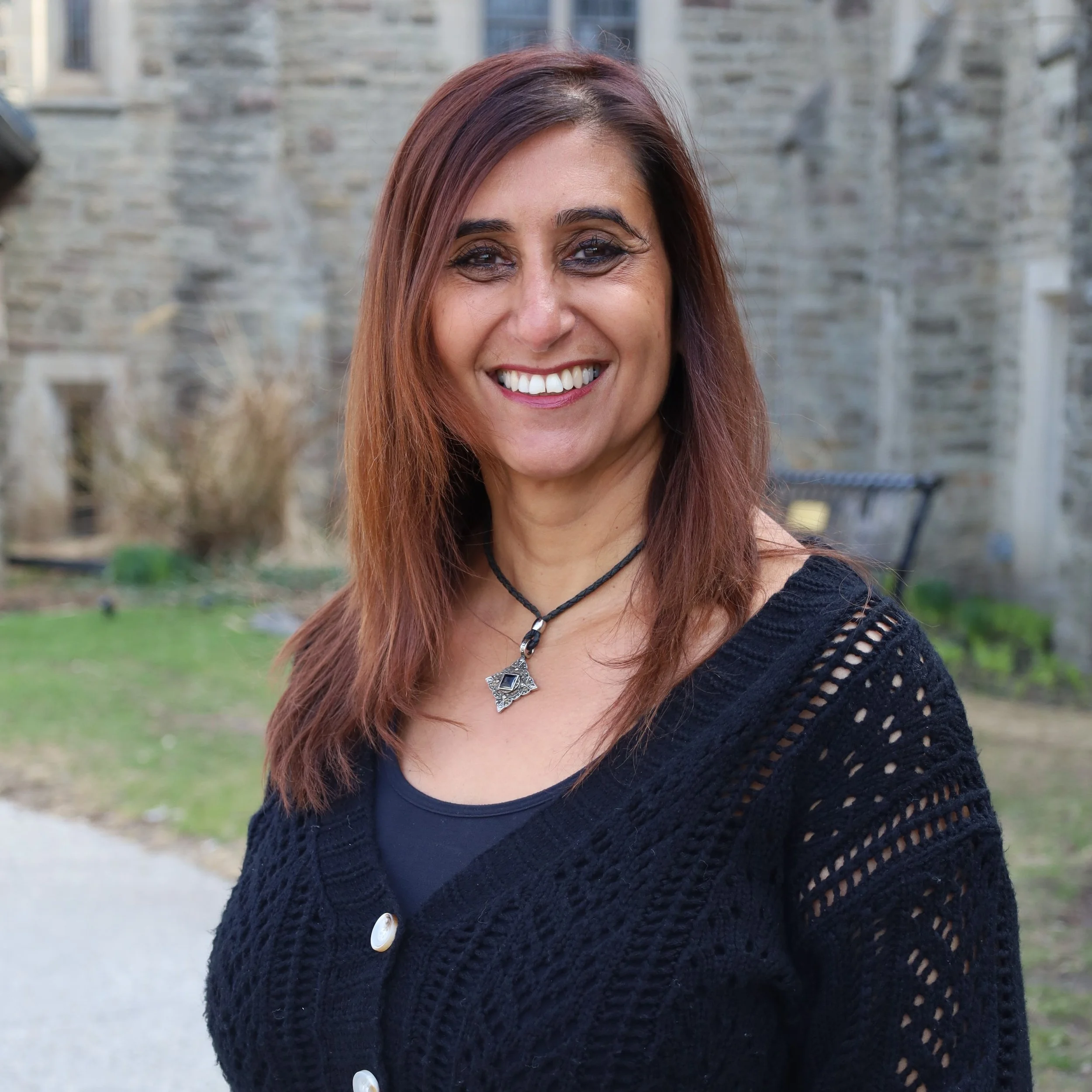 A woman with long brown hair, outdoors in front of green plants and a brick wall.