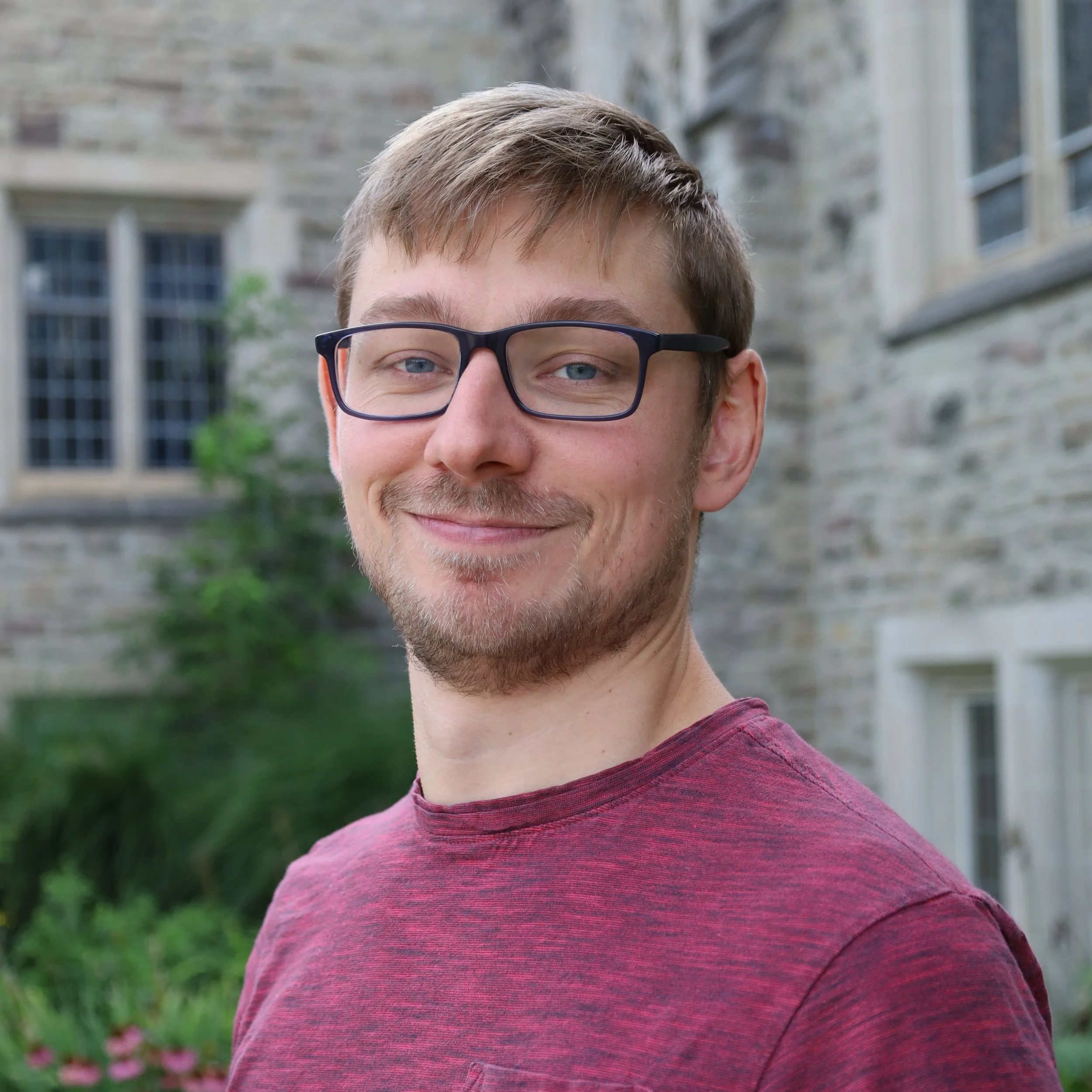 A young man with glasses and a beard smiling outdoors with a stone building and greenery in the background.